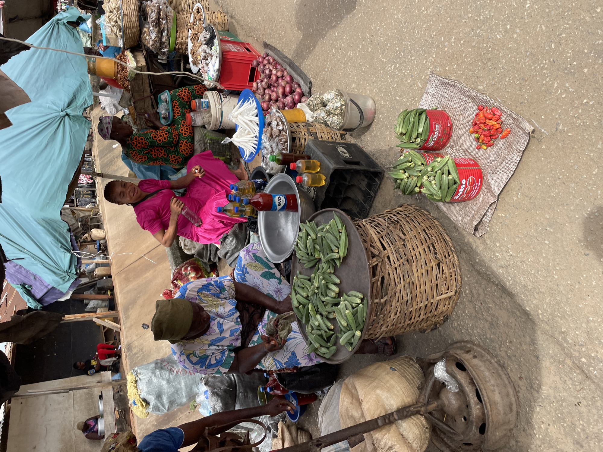 This media file captures a vibrant outdoor market scene, bustling with activity under what appears to be a bright, sunny day. The ground is a mix of concrete and packed earth, typical of an open-air market.
The primary subjects are several women, likely vendors, engaged in selling produce. In the foreground, a woman wearing a bright pink dress sits, holding a water bottle and smiling directly at the camera, suggesting a friendly interaction. Beside her, another woman in a patterned dress is seated next to a large basket overflowing with okra. A light blue umbrella is partially open, providing shade over part of the market stall.
Numerous goods are displayed for sale. Prominently featured are large quantities of fresh okra, presented in woven baskets and red cans that bear the brand name "SALSO" (likely tomato paste cans repurposed for produce). A pile of red onions is visible on the ground, and a small heap of vibrant red and orange peppers sits on a burlap mat. Various bottles, some containing red sauce or oil and others yellow oil, are arranged on a crate. Other market items include various goods in wicker baskets, metal bowls, plastic containers, and bags.
In the background, more market stalls and structures can be seen, with other individuals moving around. The overall scene conveys a lively commercial environment. The bright lighting indicates it is daytime. The specific types of produce and market setup suggest a location in a warm climate, possibly in Africa.