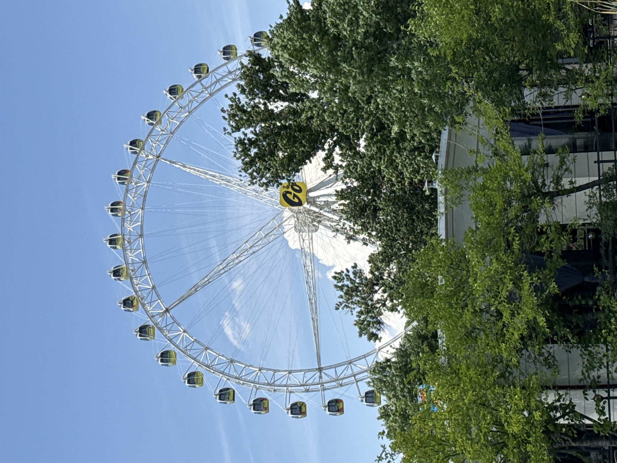 The image shows the Ferris wheel in Tashkentland, a theme park in Tashkent, Uzbekistan. The Ferris wheel is predominantly white and has a central yellow sign with black lettering. The cabins attached to the wheel are dark colored. The sky behind and around the Ferris wheel is blue with scattered white clouds. Lush green trees partially obscure the right side of the wheel and background, and a building can be seen obscured behind the trees.