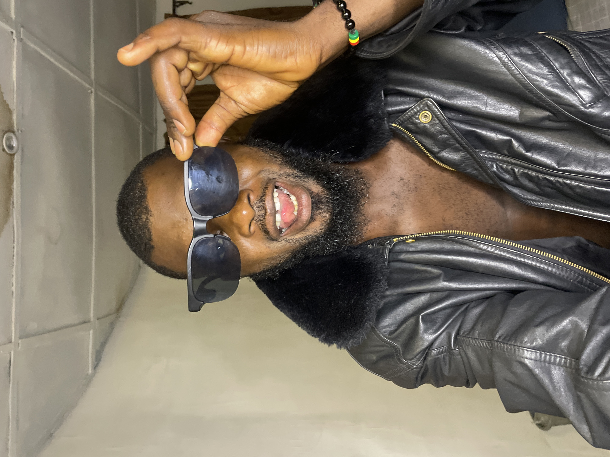A bearded man is wearing a black leather jacket with a fur collar. He has sunglasses in his hand. A black bracelet with Rasta colors is on his wrist. The background is a light-colored wall with a slight sheen. The image could be in Jos, Nigeria, but there are no details to confirm the location.
