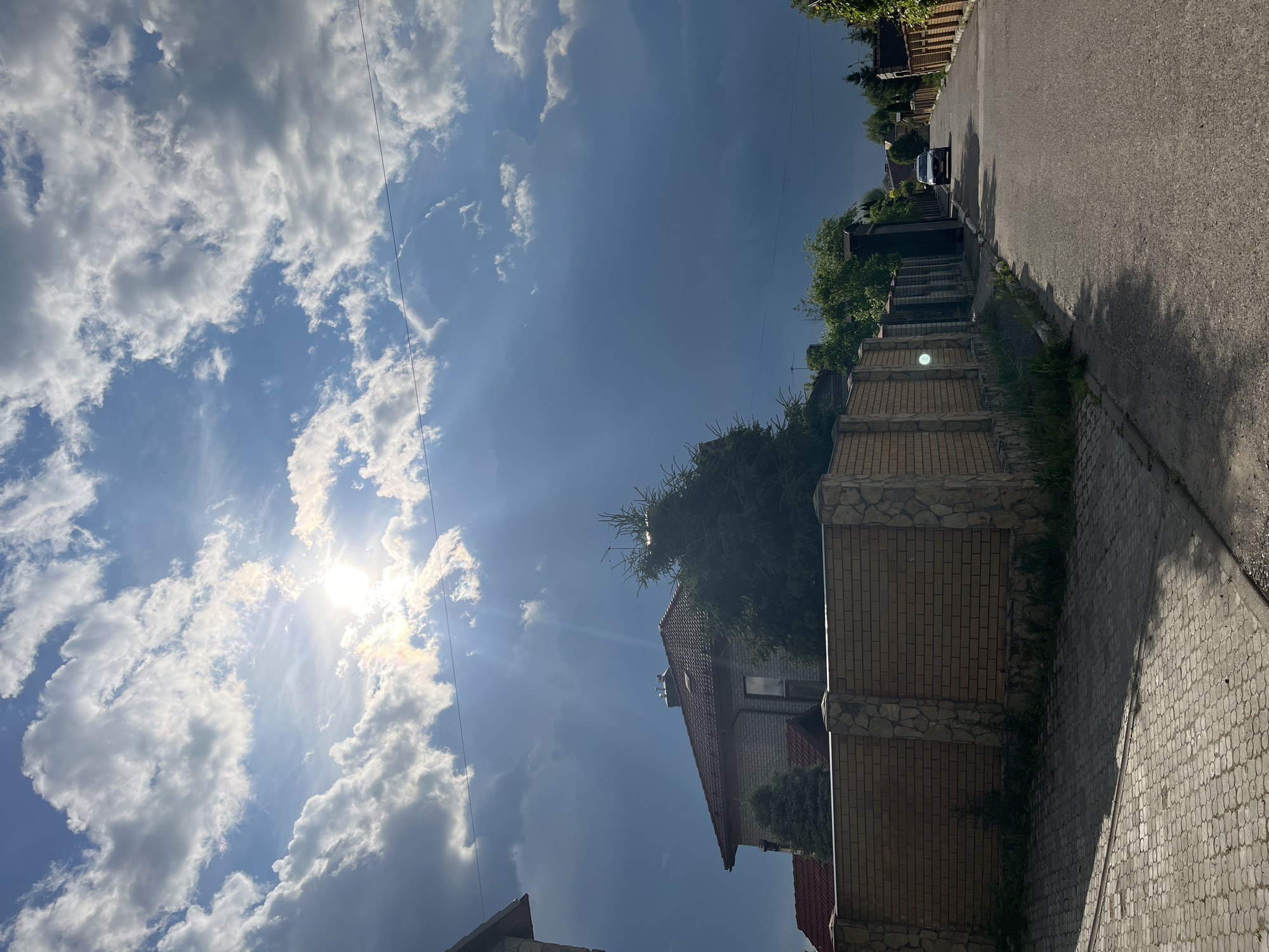 The image depicts a residential street in Orekhovka, Russia, under a partly cloudy sky with the sun shining through. The foreground features a paved walkway and a brick fence with stone accents. Beyond the fence, a house with a red tile roof is visible, surrounded by evergreen trees. A paved road runs alongside the walkway, with a single car driving in the distance. The sky is a mix of bright blue and darker gray clouds, with sunbeams visible through the clouds. The overall scene suggests a suburban environment during daylight hours.