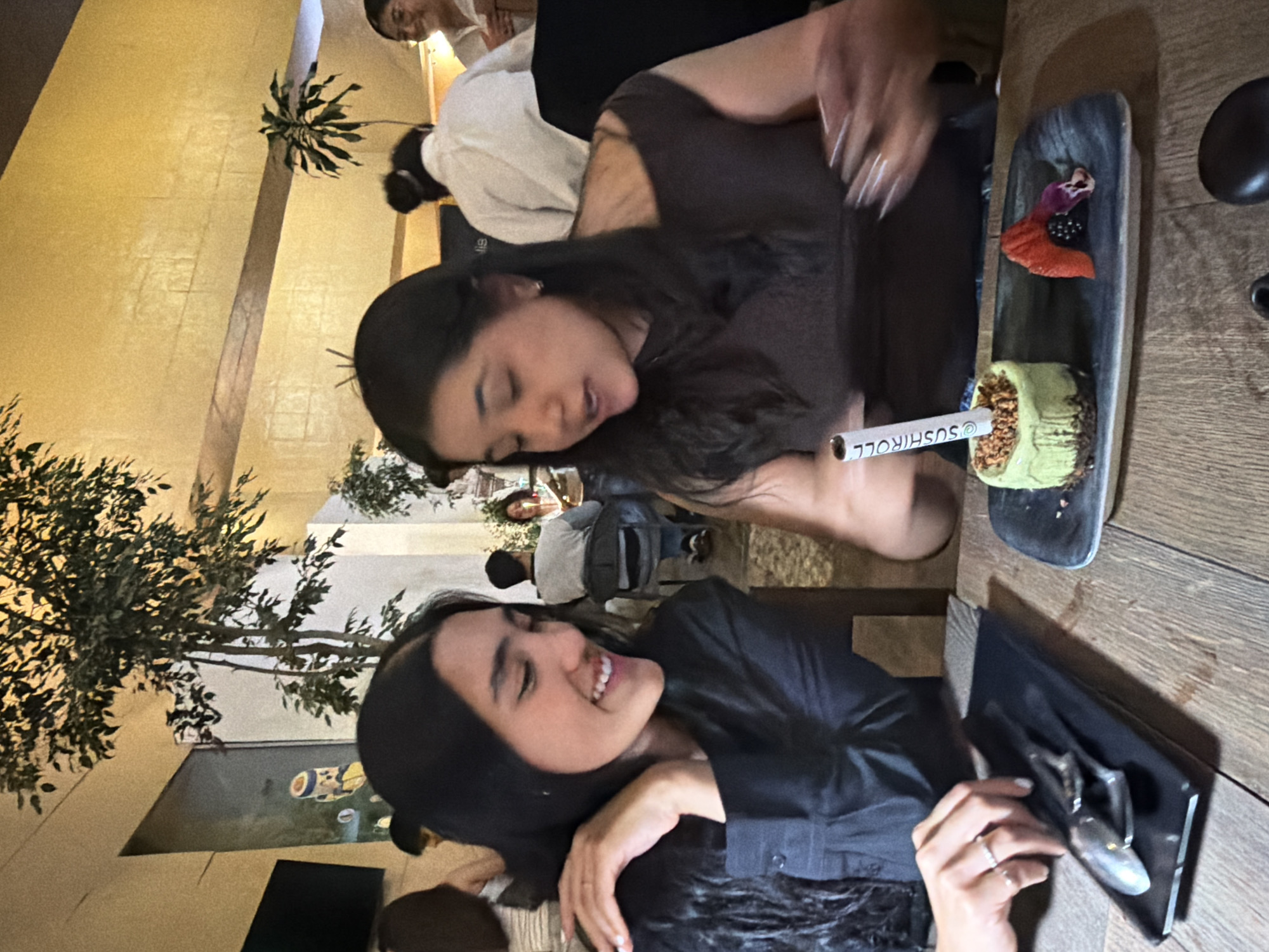 Two women are at a wooden table in a restaurant, likely in Puebla, Mexico, based on the decor and ambiance. The woman on the left wears a black shirt and smiles. The woman on the right wears a brown shirt. On the table is a plate with food, including what appears to be a sushi roll with a "Sushiroll" wrapper, and a flower garnish. A serving spoon or fork is present. The restaurant has indoor plants. Other patrons are visible in the background. The lighting is warm and dim.