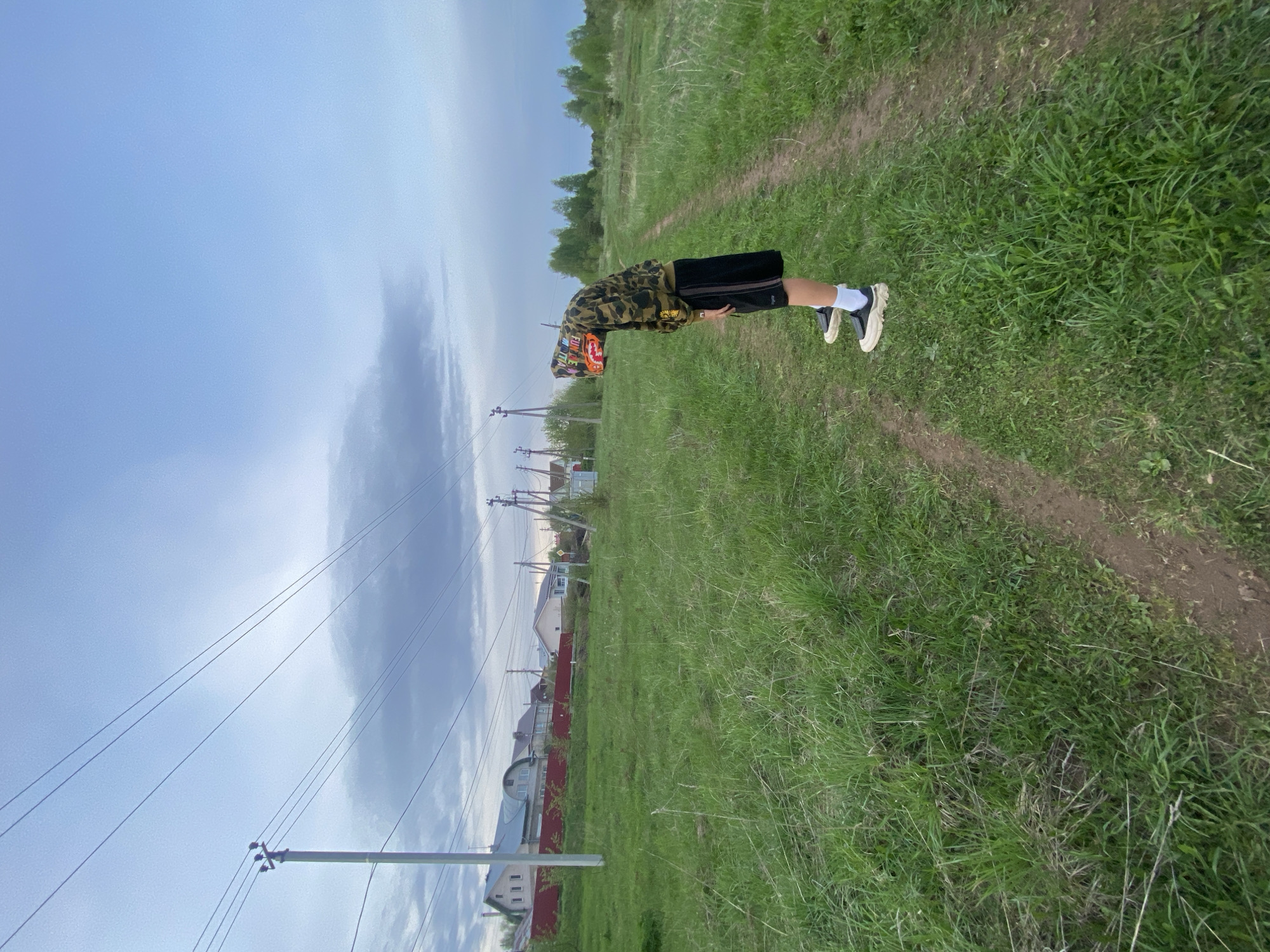 A person in a camouflage hoodie and black shorts stands on a grassy path in Прикамский, Russia. The path is bordered by tall grass and a red fence. In the background are houses and power lines under a cloudy sky. The person is bending forward, looking down at the ground.