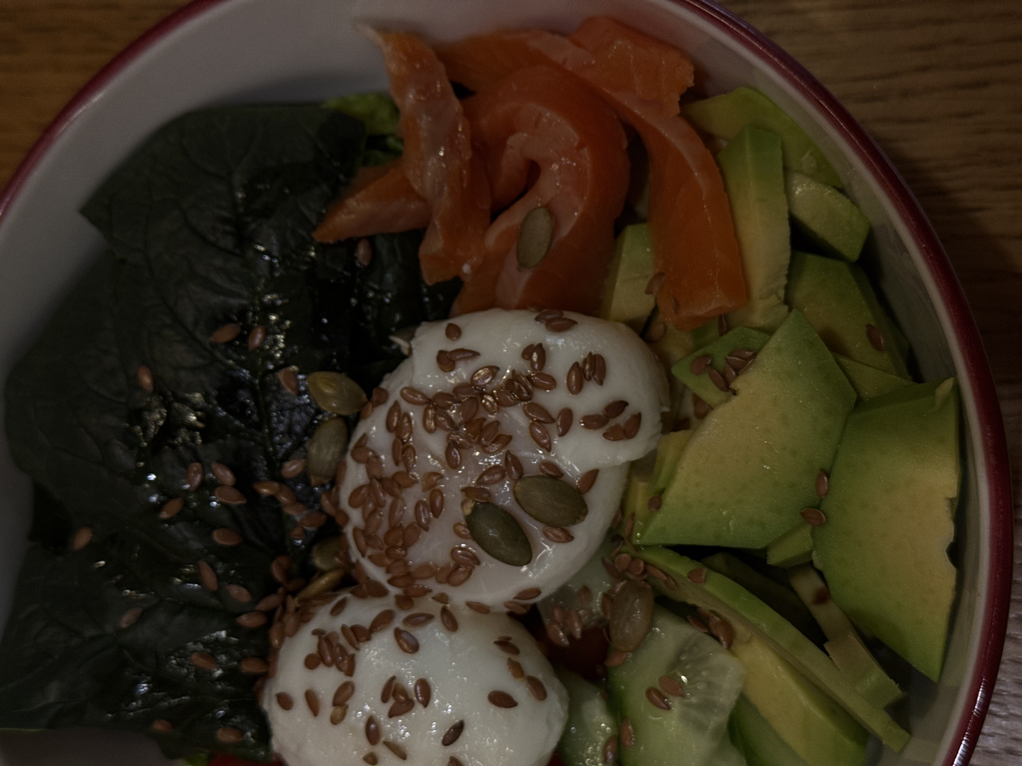 A close-up, overhead shot depicts a salad bowl in a restaurant, possibly in Krasnoyarsk, Russia. The bowl contains what appears to be dark leafy greens, slices of raw salmon, wedges of avocado, cucumber slices, two poached eggs, flax seeds, and pumpkin seeds. The ingredients are arranged in distinct sections within the bowl. The rim of the bowl is visible and features a red accent. The background suggests a wooden surface.