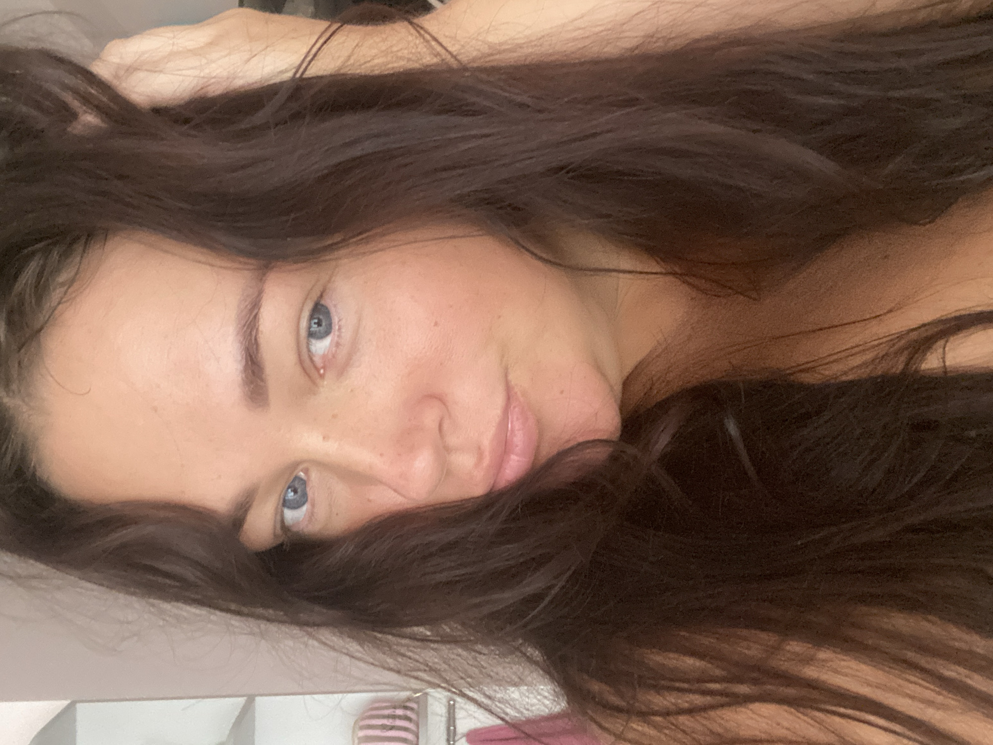 The image shows a close-up selfie of a woman with long, dark brown hair and blue eyes. Her hair is falling around her face, and she is touching her head with her right hand. Her head is tilted slightly to the left, and she is looking directly at the camera with a slight smile. The background appears to be a wall with some items on a shelf, but it is out of focus. There is no indication of the location in Sochi, Russia.
