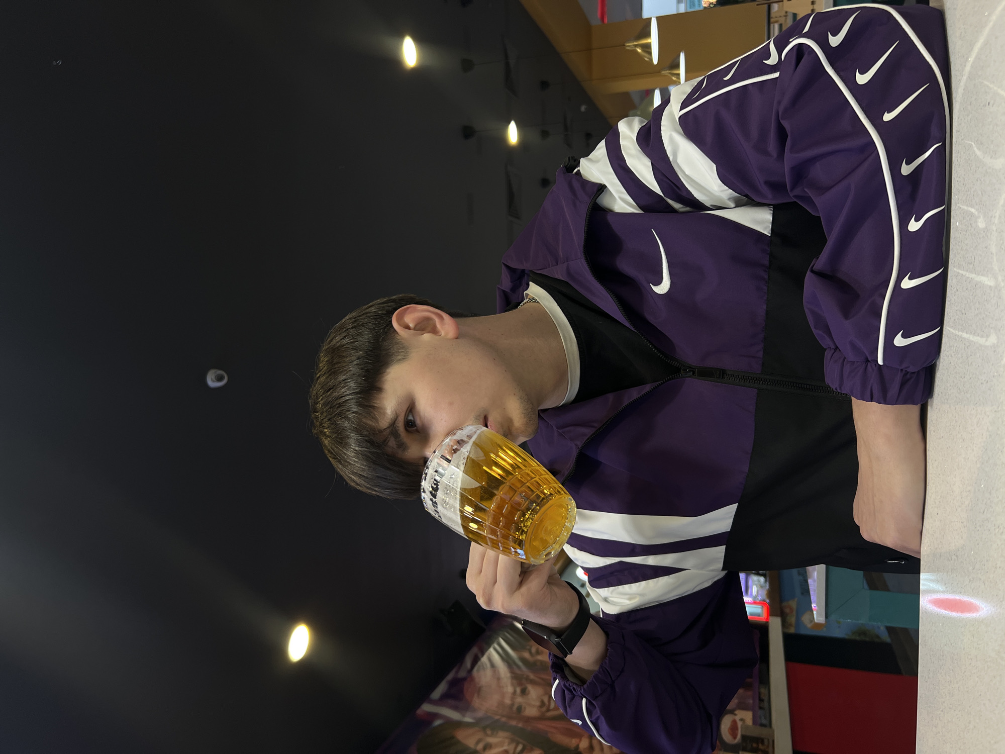 A young man is drinking beer from a glass mug. He is wearing a purple and white Nike jacket. The scene is lit by hanging light fixtures. The background is dark.