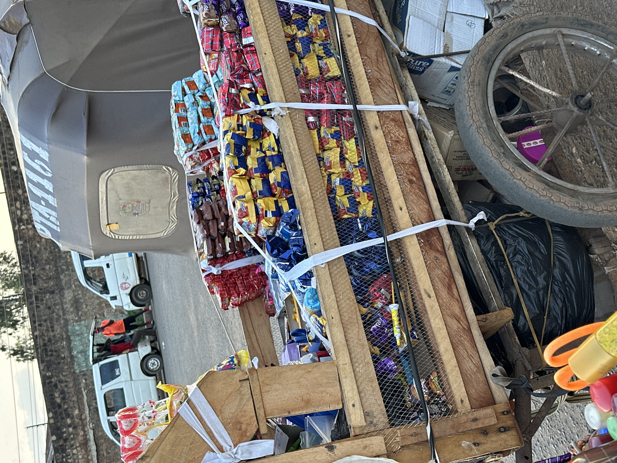 This image captures a street scene in Bukuru, Nigeria, focusing on a vendor's cart laden with an assortment of goods. The cart itself is a rustic wooden structure, possibly a modified bicycle or motorcycle trailer, with a large, worn wheel visible on the right. It is packed with various items, predominantly snacks and candies. Stacks of colorful wrappers, some in shades of red, blue, and yellow, suggest a variety of confectionery products. These are neatly arranged in wooden crates and secured with white straps.
A prominent object on the cart is a large black plastic bag, possibly containing more merchandise or supplies, tied with rope. Beside it, a bright orange object with a loop, likely a part of a toy or promotional item, is visible. Further items, including small bottles and colorful plastic containers, are arranged at the bottom right of the cart, hinting at the diverse nature of the vendor's offerings.
In the background, a light-colored vehicle, possibly a minivan or utility truck, is parked, with people visible inside. One person in an orange shirt appears to be leaning out. Another similar vehicle is seen further down the road. The presence of these vehicles suggests a roadside or urban setting. The overall lighting indicates it could be daytime, possibly late afternoon or early morning, given the angle of the light. The scene appears to be bustling with activity, typical of a market or roadside vending area. The condition of the cart and the items suggest a hardworking vendor operating in a practical, everyday environment.