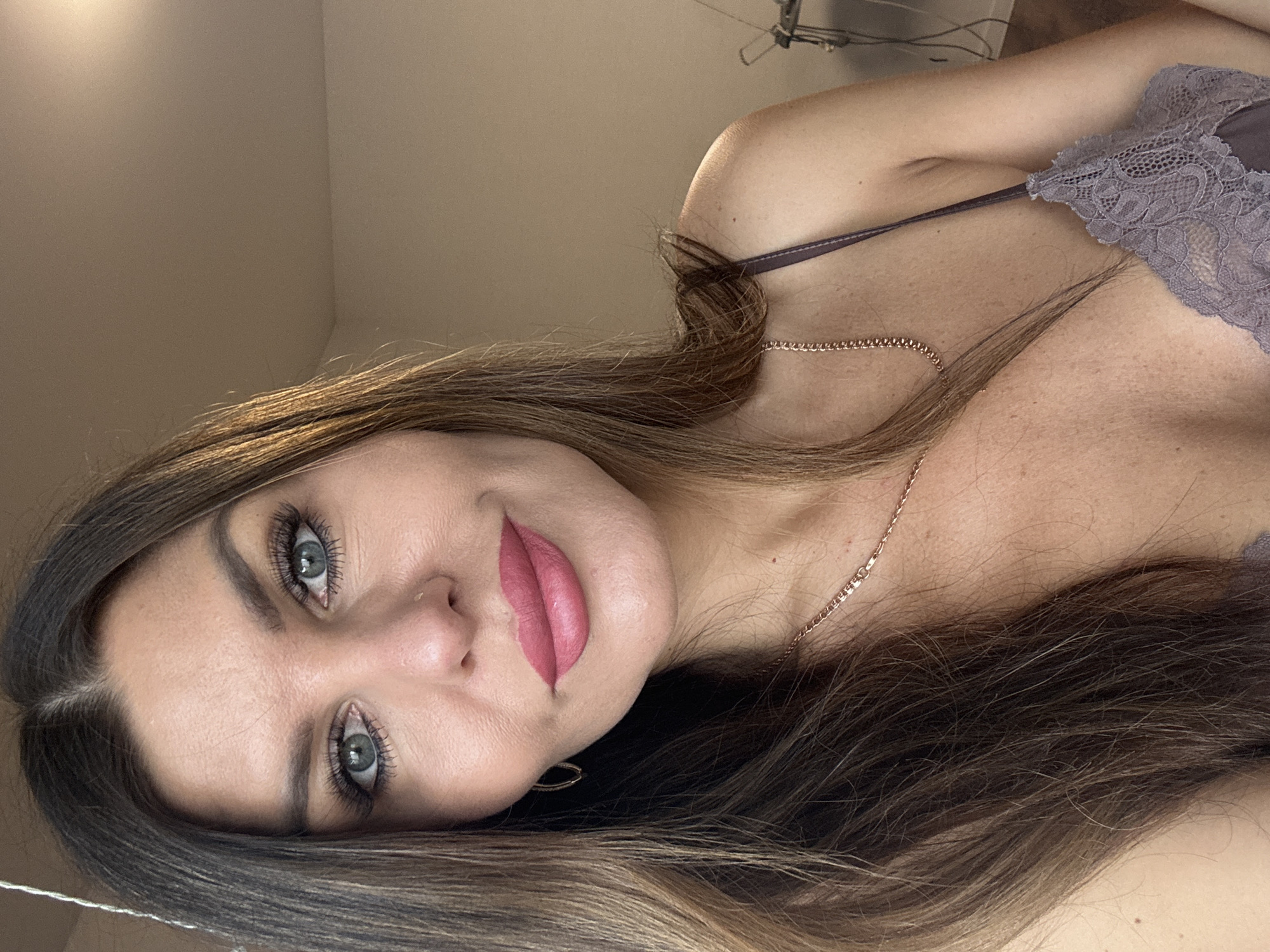 A close-up portrait shows a woman from the chest up, reclining with her head tilted towards the camera. Her long, dark brown hair, highlighted with lighter caramel or blonde tones, frames her face and cascades across her chest. She has striking blue-green eyes, enhanced by long, dark eyelashes and well-defined eyebrows. Her full lips are adorned with a matte rose-pink lipstick, and she presents a subtle, closed-mouth smile. She is wearing a delicate, grayish-purple or lilac lace bralette or camisole with thin straps. A thin, gold-colored chain necklace rests on her décolletage. The background consists of a plain, light-colored wall, indicating an indoor setting. Some wires and a fixture are faintly visible in the upper right background. The woman maintains a direct and confident gaze. The location is Chaikovsky, Russia. No text is visible.