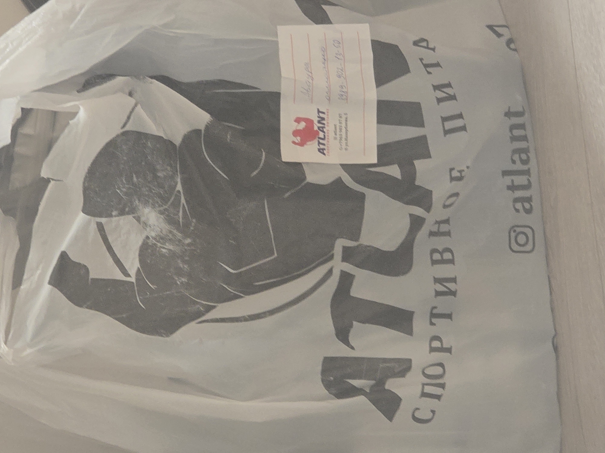 The image shows a white plastic bag with a dark grey graphic of a muscular man. The word "ATLANT" is printed prominently in large grey letters across the bag, with smaller grey text below reading "СПОРТИВНОЕ ПИТА". There is also a small white sticker with a red logo and handwriting in Russian. The sticker has the "ATLANT" logo, a website address, and a phone number. Below the main "ATLANT" text on the bag, there is an Instagram logo and the word "atlant". The handwritten text on the sticker appears to be a name and some numbers. The environment is indoors, with a light-colored floor visible at the bottom of the image. There are no people or specific landmarks visible that would indicate the city of Avtury, Russia.