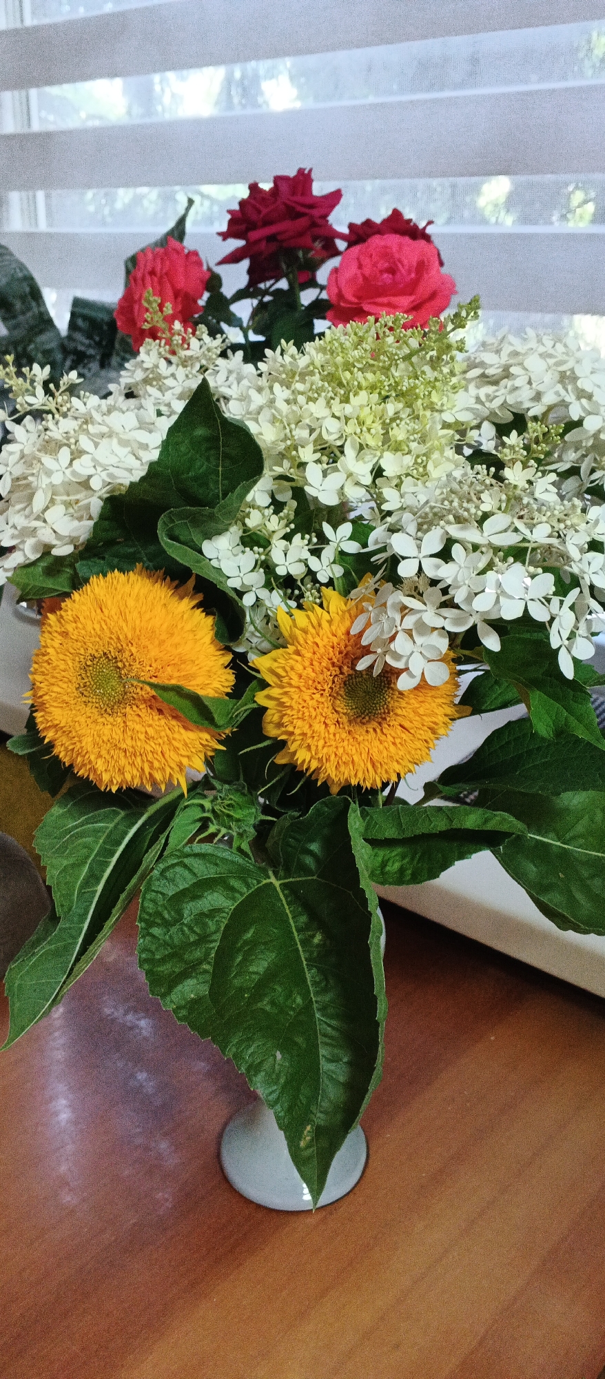 A close-up, vertical shot captures a bouquet of flowers resting on a wooden table. The arrangement includes two fluffy, bright yellow sunflowers in the foreground, their centers a vibrant green. Behind them, delicate clusters of small white flowers, possibly hydrangeas, are interspersed with lush green leaves and stems. Two deep red roses and a bright pink rose add a touch of color towards the top of the bouquet. The flowers are contained within a light-colored vase, the bottom of which is visible on the wooden surface. The background is out of focus, showing what appears to be white blinds on a window, with glimpses of greenery beyond. The lighting suggests it is daytime. The location is not specified within the image.