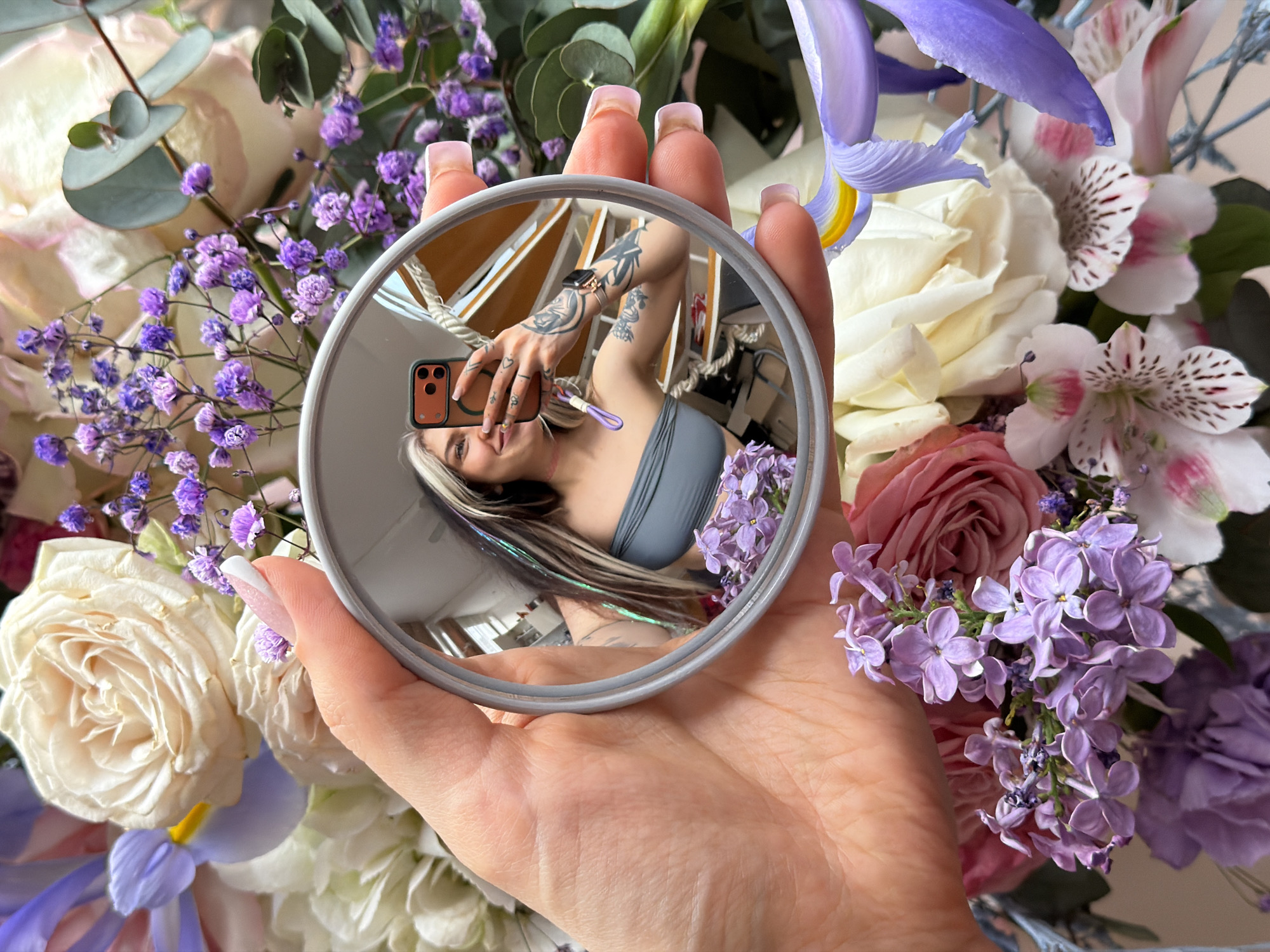A woman's hand holds a circular mirror against a background of flowers. The mirror reflects the woman, who is taking a picture of herself with a brown iPhone. She is wearing a gray top and has several tattoos on her arm. The reflection reveals more of the interior behind her, including a table and other objects. The flowers include roses, irises, and other small purple blossoms. The woman is not in Budva, Montenegro.