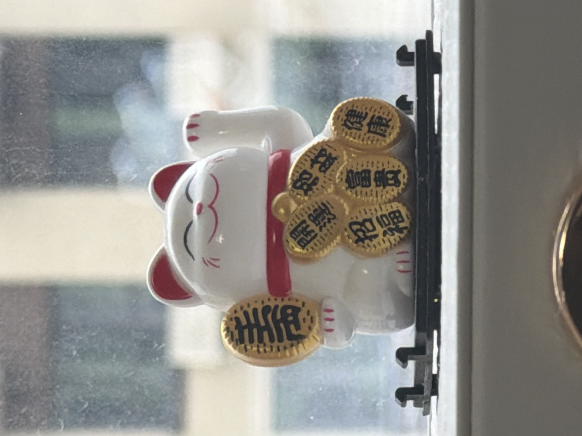 A white ceramic Maneki-neko, or lucky cat figurine, sits on a black plastic stand against a blurred background of a window. The cat has a red collar and is holding a gold coin in its left paw. Several other gold coins are arranged in a semi-circle around the cat's body, each inscribed with Chinese characters. The figurine appears to be positioned on a windowsill. No specific location in Kazan, Russia, can be determined from the image.