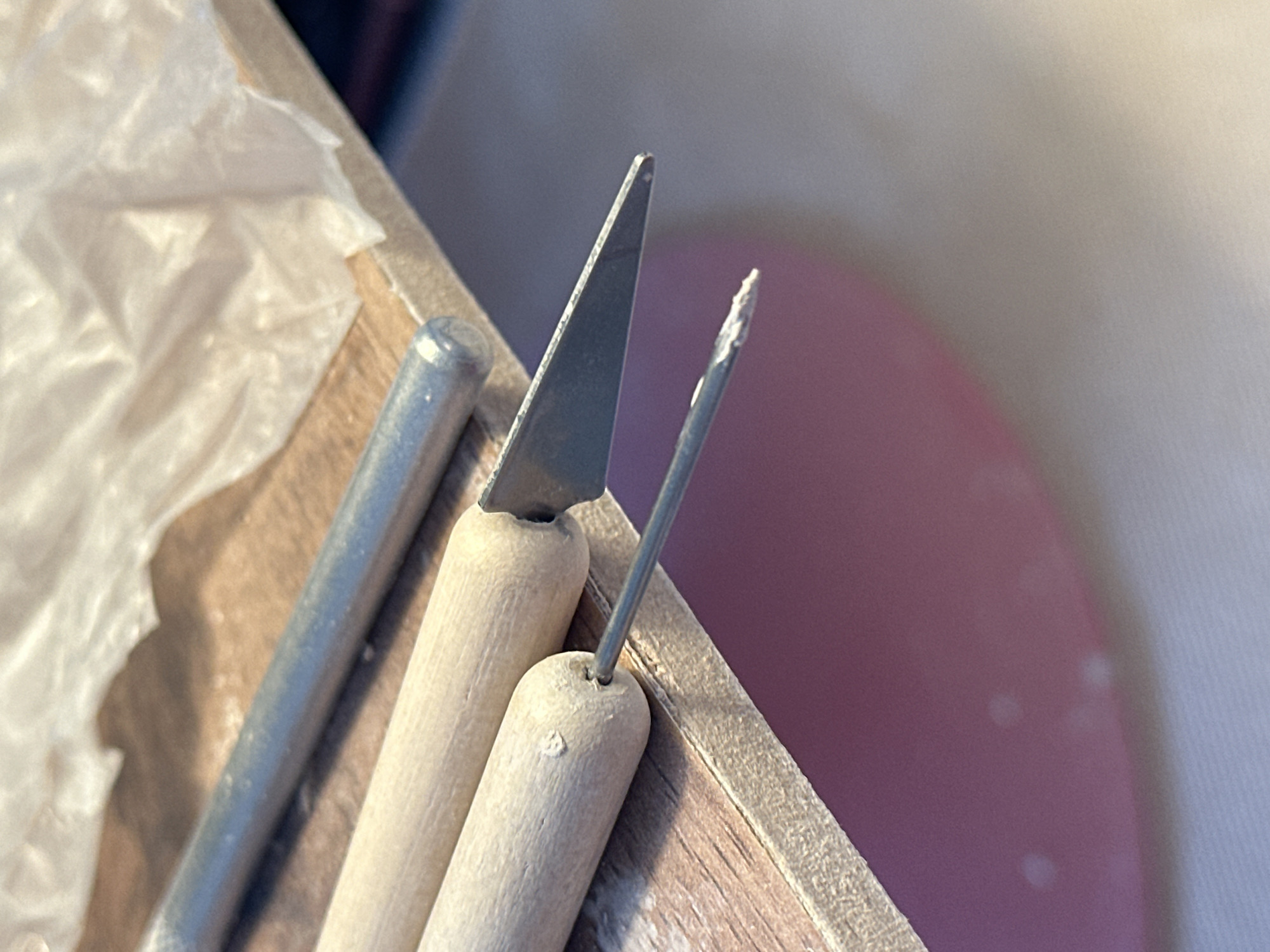 The image shows a close-up of sculpting tools on a wooden surface. Two wooden-handled tools are visible, one with a pointed metal tip and another with a broader, angled metal blade. A third, unadorned metal tool lies above them. The background includes a crumpled piece of translucent material and a blurred pink surface below. There is no indication of the city of Nevinnomyssk, Russia, or any specific location. The image is a still life composition of art supplies.