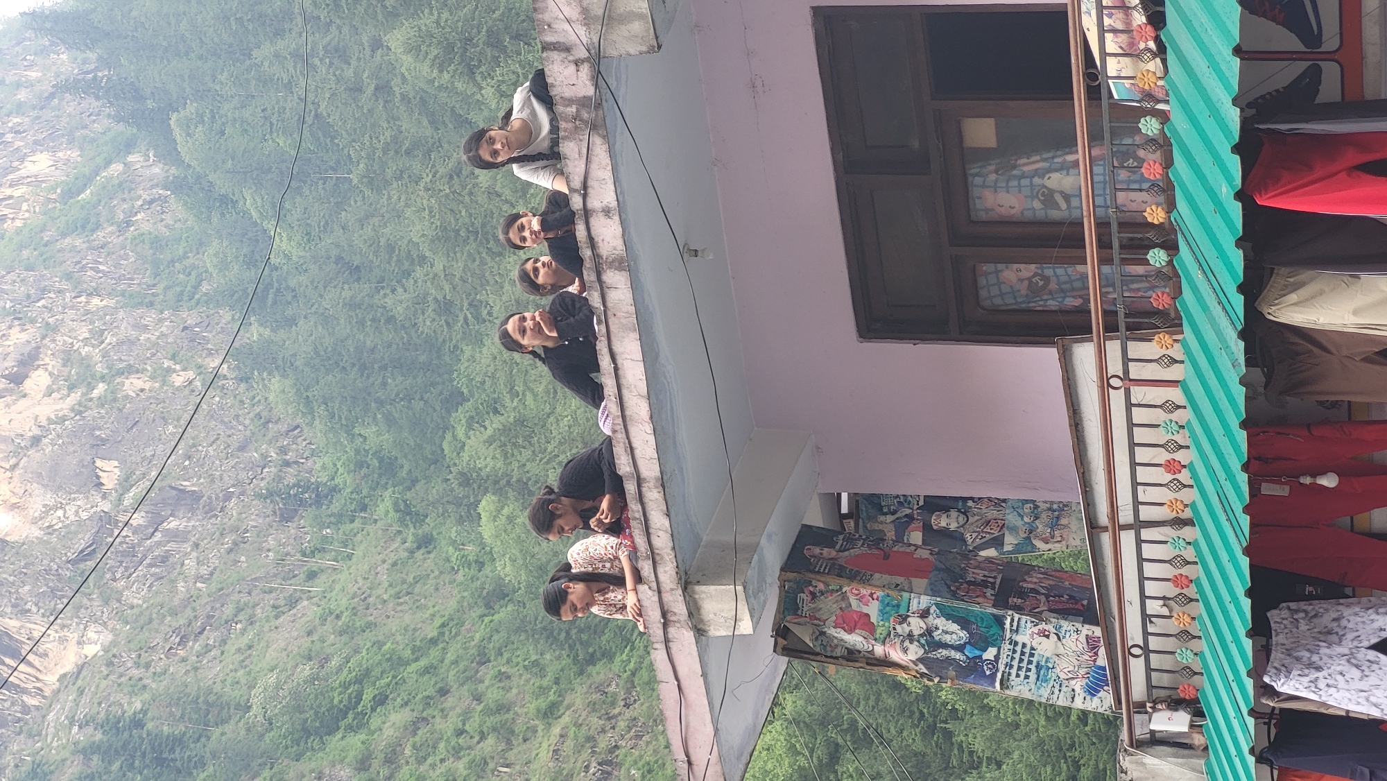 This image, set in Manikaran, India, captures a scene from a building overlooking a mountainous landscape. In the foreground, a group of approximately seven women and girls are leaning over the edge of a flat rooftop or balcony, gazing towards the left side of the frame. Their expressions are primarily curious and observant, with some smiling, suggesting engagement with what they are viewing. They are dressed in a mix of casual wear, including white tops, dark sweaters, and patterned blouses.
The building itself features a light pink facade with dark-framed windows visible below the balcony level. Directly below the balcony and extending to the right, a vibrant green corrugated metal roof covers a lower section, likely a shop. Various clothes, including red pants, beige trousers, and patterned shirts, hang on display near this roof, indicating a retail space. A decorative metal railing with flower-like patterns in green, orange, and yellow adds a touch of adornment to the building. On the wall beneath the balcony, several posters with vibrant imagery and depictions of people are visible, though any specific text is illegible. A single light bulb hangs from a wire near the shop area.
Beyond the urban elements, a sprawling natural environment dominates the background. Dense green forests cover the slopes of mountains, which recede into the distance. A large, rocky mountain face is visible further left, its contours partially softened by atmospheric haze or mist. Utility wires stretch across the scene, connecting elements within the valley. The diffused, soft lighting suggests it is daytime, possibly morning or late afternoon, under an overcast or hazy sky, creating a gentle illumination without harsh shadows. The overall impression is a blend of daily life in a bustling area set against a majestic natural backdrop.