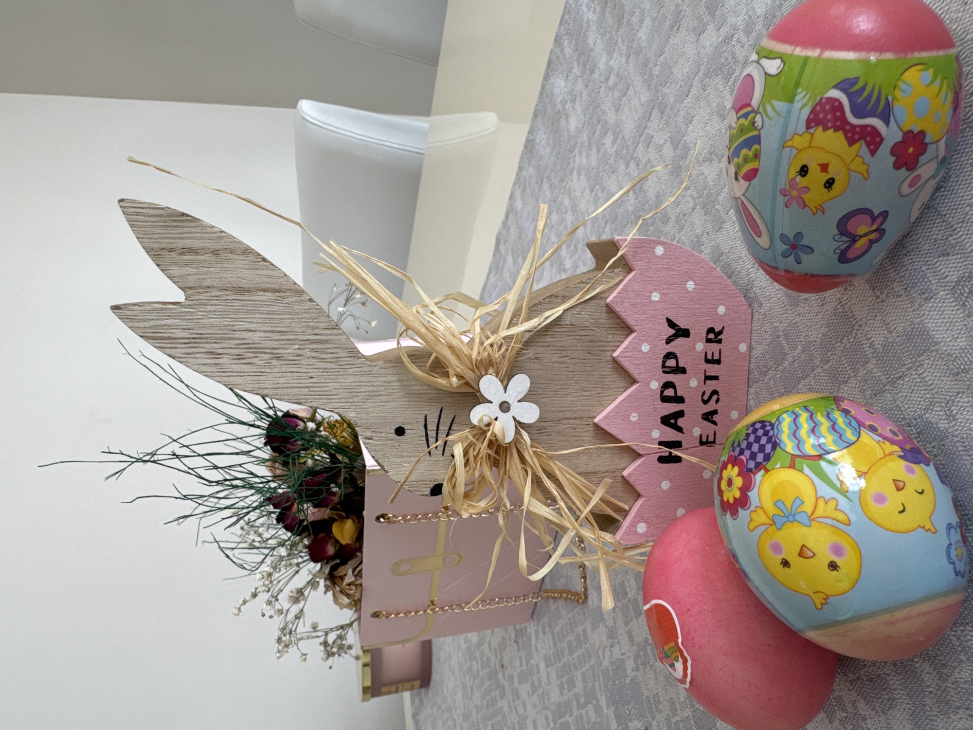 The image displays a close-up of Easter decorations on a gray textured surface. A wooden bunny cutout with a pink "Happy Easter" tag is the central element. The bunny is adorned with dried flowers and straw. Next to the bunny are three Easter eggs, two with chick designs and one solid pink. The background includes a partial view of a white chair and a plain white wall. The location and people involved are unknown. There is no information to suggest this image was taken in Varna, Bulgaria.