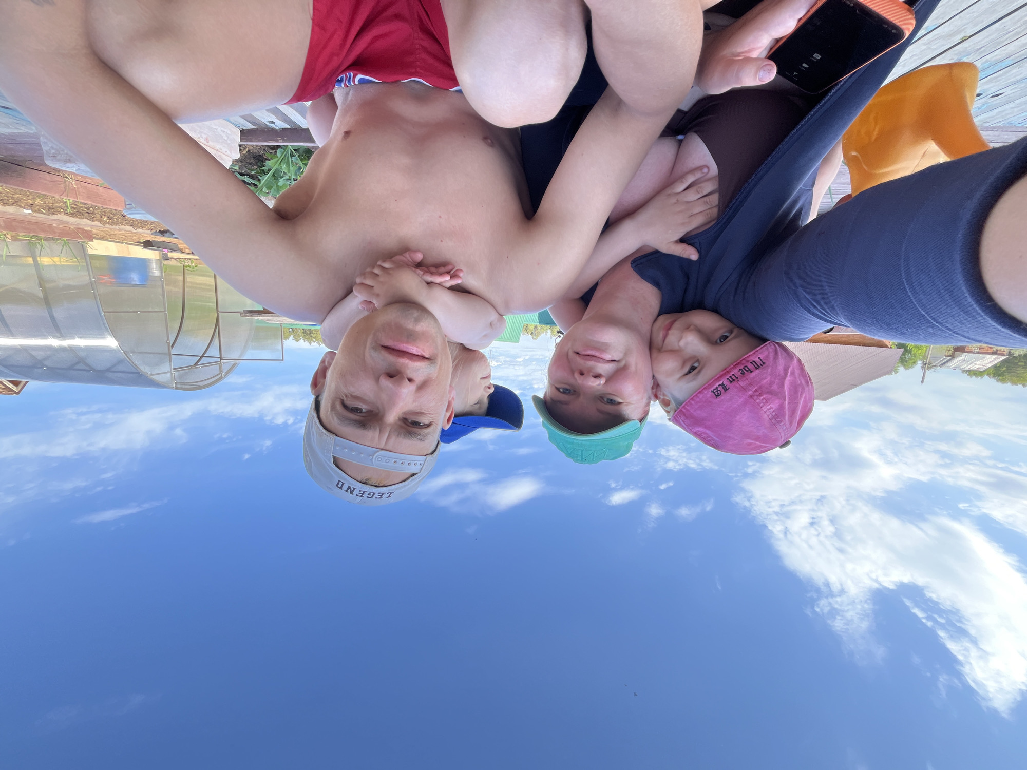 The image shows an upside-down selfie taken by a man in a grey baseball cap with the word "LEGEND" on it. He is shirtless and wearing red swim trunks. He is holding a small child with fair skin and a blue cap. Next to him are two children, one with a turquoise cap and the other with a pink cap that reads "I'll be in LA." A woman with a phone in hand is beside the child with the pink cap. They are positioned in front of a clear blue sky with light clouds. In the background, slightly out of focus, is a greenhouse, some wooden planks, a building, and a yellow object, possibly a toy. Based on the information and visual clues, the location is in the city of Chaikovsky, Russia.