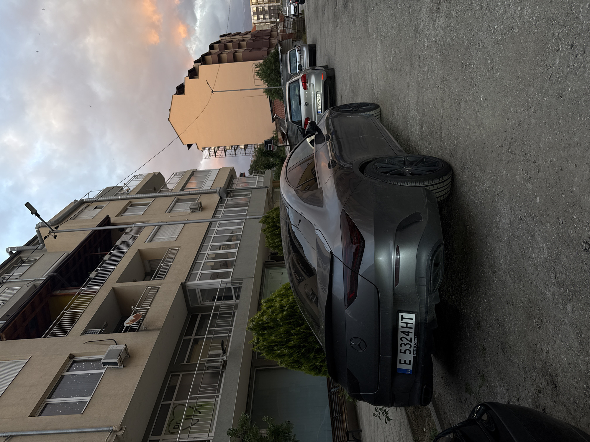 The image shows a street scene in Blagoevgrad, Bulgaria. Two vehicles are parked on the side of the road: a dark grey Mercedes-Benz with license plate "E 5324 HT" and a light grey Volkswagen with license plate "E 9526 HH". Buildings line the street, with one showing extensive exterior ladders and access platforms. The sky exhibits a sunset glow, with orange and grey hues.
