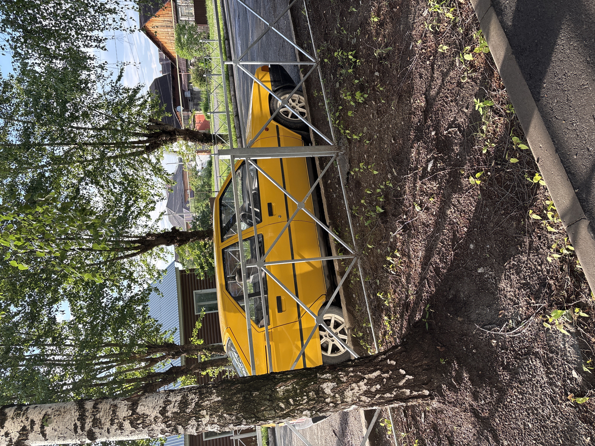 A yellow, four-door sedan is parked behind a metal fence. The car appears to be older, with visible wear. The scene is located in the city of Rezh, Russia, as indicated by the presence of typical Russian dacha-style houses and wooden fences in the background. The area around the car is covered in dirt and sparse vegetation, with a large tree on the left side of the frame. The sky is clear and blue, suggesting it is daytime. There are no people visible in the image. The lighting suggests it is late afternoon or early morning, with long shadows cast by the trees.