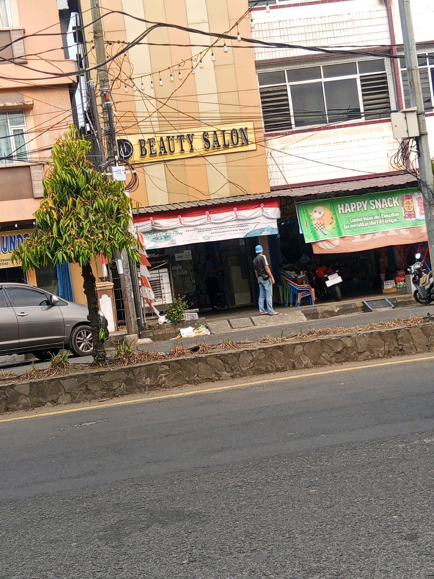 The image shows a street scene in Lubuklinggau, Indonesia. In the foreground is a two-lane asphalt road with a yellow dividing line. A low stone wall borders the sidewalk, which is paved with concrete and lined with sparse vegetation.
On the sidewalk, a gray sedan is parked on the left. To the right of the car is a slender tree with dense green foliage. Further to the right, a building with a facade of horizontally striped panels in shades of beige and orange houses several businesses. A prominent sign with gold lettering reads "BEAUTY SALON." Below this sign, a red and white banner is partially visible.
Adjacent to the beauty salon is another storefront with a large green banner that says "HAPPY SNACK" and displays an image of a cartoonish shrimp. This banner also features text in Indonesian, which appears to be promotional. A man wearing a blue cap, black t-shirt, and blue jeans stands on the sidewalk, facing the stores. Several colorful plastic chairs are stacked near the entrance of one of the establishments. A motorcycle is parked in front of the "Happy Snack" store. A second, darker motorcycle is parked to its right. Overhead, a complex network of electrical wires and a string of decorative lights are visible. The building to the right of the beauty salon has a white tiled facade and large windows with horizontal blinds.
