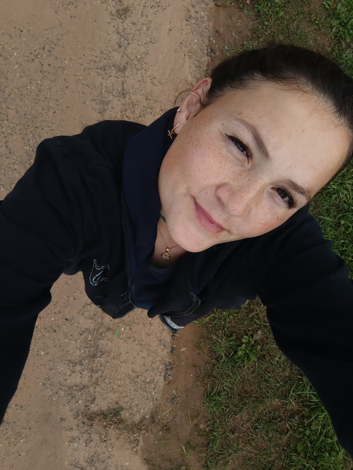 A high-angle, outdoor selfie shows a woman with dark hair pulled back, a black hoodie, and a gold necklace. She has freckles across her nose and cheeks and is smiling. The background consists of dirt and grass. The image does not contain any elements that indicate the location as Karsha, Russia.