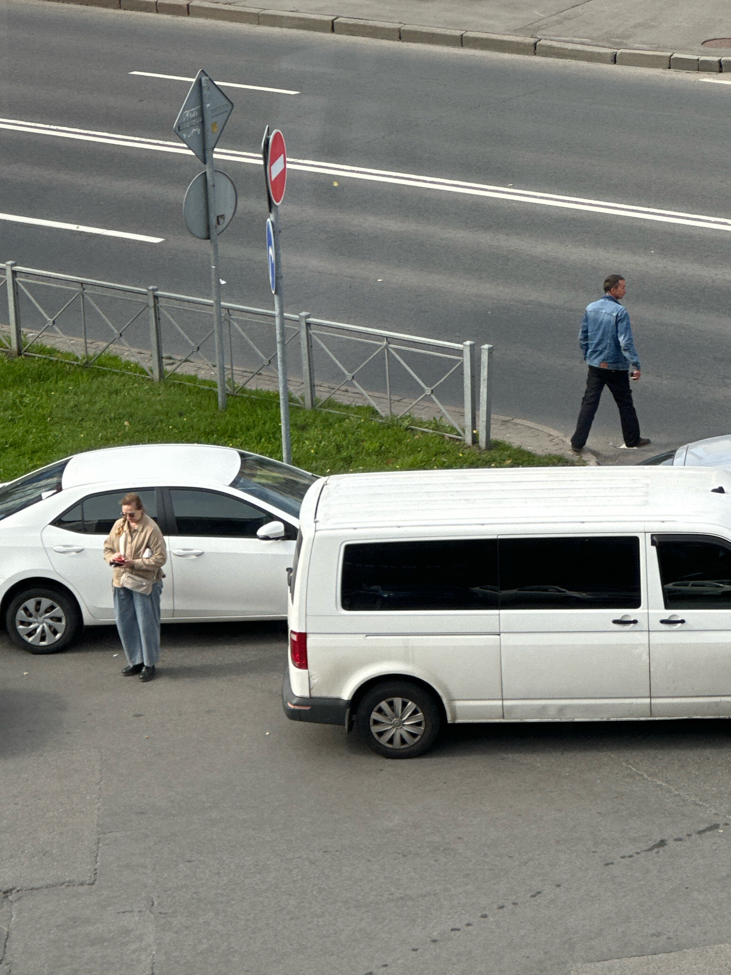 A street scene in Saint Petersburg, Russia, features two white vehicles, a sedan and a van, parked on a paved road. A woman in a light-colored jacket and jeans stands next to the sedan, looking at her phone. A man in a denim jacket and dark pants walks away from the viewer on the sidewalk. Traffic signs are visible on poles along the road. Green grass and a metal fence line the side of the road. The asphalt is marked with white lane lines.