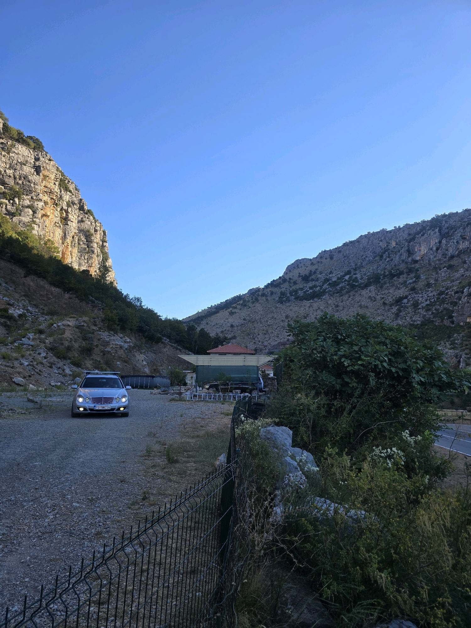 A silver Mercedes-Benz E-Class sedan is parked on a gravel lot in a canyon near Podgorica, Montenegro. The car, with its headlights on, faces the camera and has a roof rack. The license plate reads "PG-NM 734". The canyon walls are steep and rocky, with sparse vegetation. In the background, a building with a red roof is visible, along with what appears to be an above-ground swimming pool. A chain-link fence runs along the right side of the frame. The sky is clear and blue, indicating daytime.