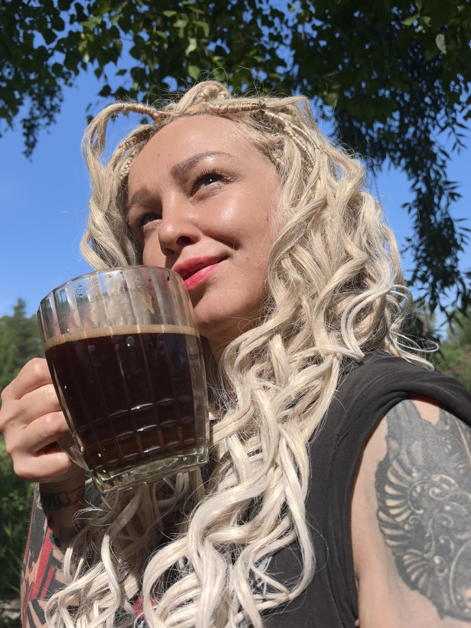 A young woman with blonde dreadlocks and red lipstick drinks from a glass mug of dark liquid. She is wearing a black top and has tattoos on her arms. She is outdoors, with green leaves and a blue sky in the background. The image was taken in Noviy, Russia.