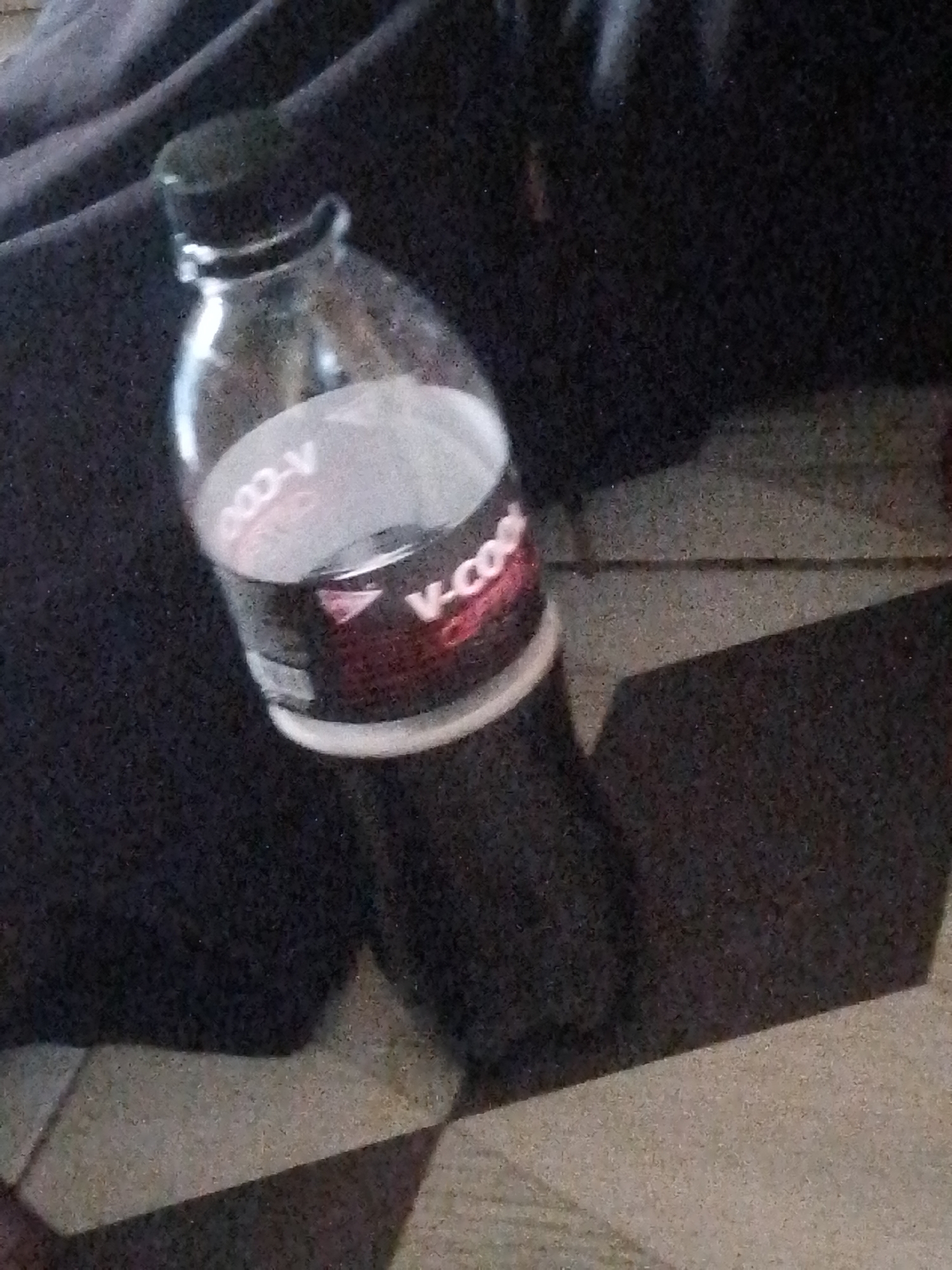 A dark-colored bottle with a black cap is positioned at an angle on a dark, textured surface. The bottle appears to contain a dark liquid. A white label is visible on the bottle, with some text in red and white lettering. The label has the words "V-COOL" in large font and "COOL" repeated. The bottle casts a shadow on the surface it rests on. The background is dark and out of focus. The floor is tiled with light and dark colored square tiles arranged in a diamond pattern.