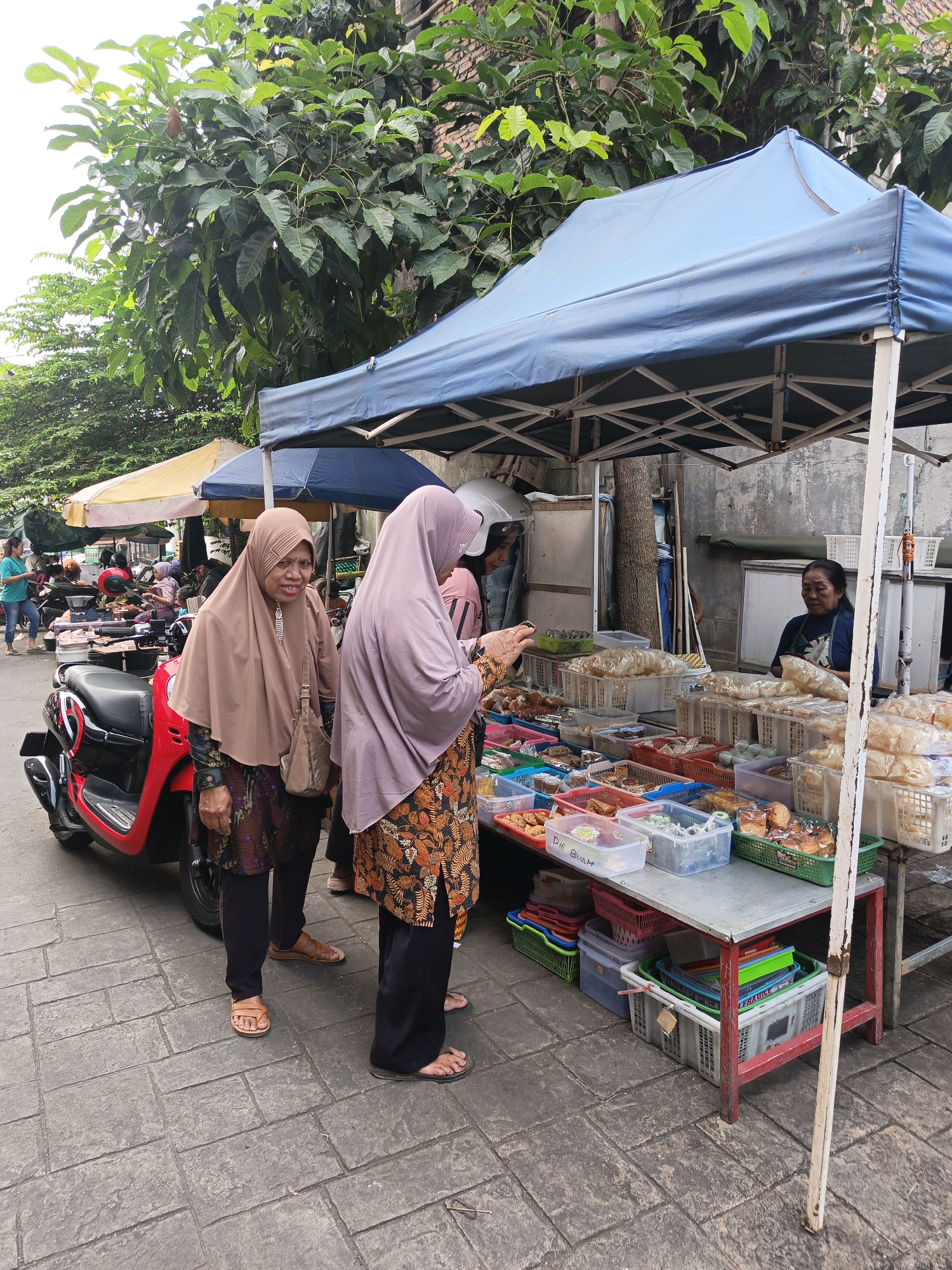 In Klaten, Indonesia, two women wearing hijabs stand at a street food stall. The woman on the left, in a light brown hijab, faces the camera with an indifferent expression, holding a small bag. The woman on the right, in a lavender hijab, faces away from the camera, examining items at the stall. The stall is covered by a blue canopy and displays various food items in plastic containers and baskets, including pastries and what appear to be snacks. A red scooter is parked to the left of the stall. In the background, other stalls, parked motorcycles, and lush green trees are visible. The ground is paved with interlocking bricks. A woman behind the counter is serving a customer.
