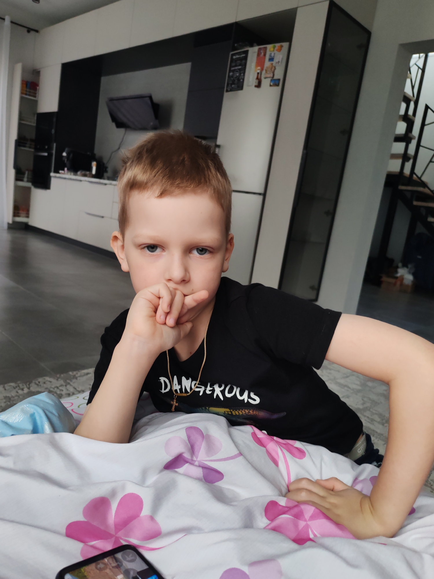 A young boy with fair skin and reddish-blond hair is lying in bed. He is wearing a black t-shirt with the word "DANGEROUS" printed on it. He has a gold chain with a cross pendant around his neck. His left hand is raised to his mouth, with his fingers touching his lips. His right hand rests on the bed next to him.
The bed has white sheets with pink and purple flower patterns. A blue object, possibly a blanket or pillow, is visible next to him on the left. A smartphone is partially visible at the bottom left corner of the frame.
The background shows a modern interior. There is a kitchen area with white cabinets and black accents. A television is mounted on the wall. In the background, there is a glimpse of a staircase with black railings.
Given the context of the prompt, it is speculated that this scene is located in a home in the city of Chaikovsky, Russia.