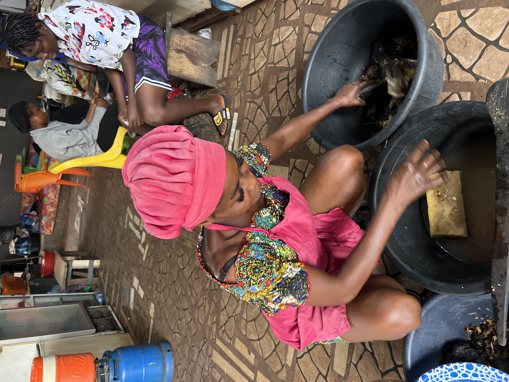 In Nkpor, Nigeria, a woman in a pink headscarf and apron is squatting down, working with two large black tubs filled with water and what appears to be raw meat or fish. One tub is closer to her and contains a yellow object, possibly a sponge or cleaning tool. Her hands are submerged in the water, suggesting she is washing or preparing the contents. The other tub, to her right, also contains similar dark items. Behind her, two other individuals are visible. One is wearing a grey garment and sitting on a bright yellow chair, while the other, wearing a white shirt with a colorful pattern and purple shorts, is seated on a wooden stool. The environment appears to be an outdoor market or processing area, with tiled flooring and various items in the background, including a gas cylinder, coolers, and stacked goods.