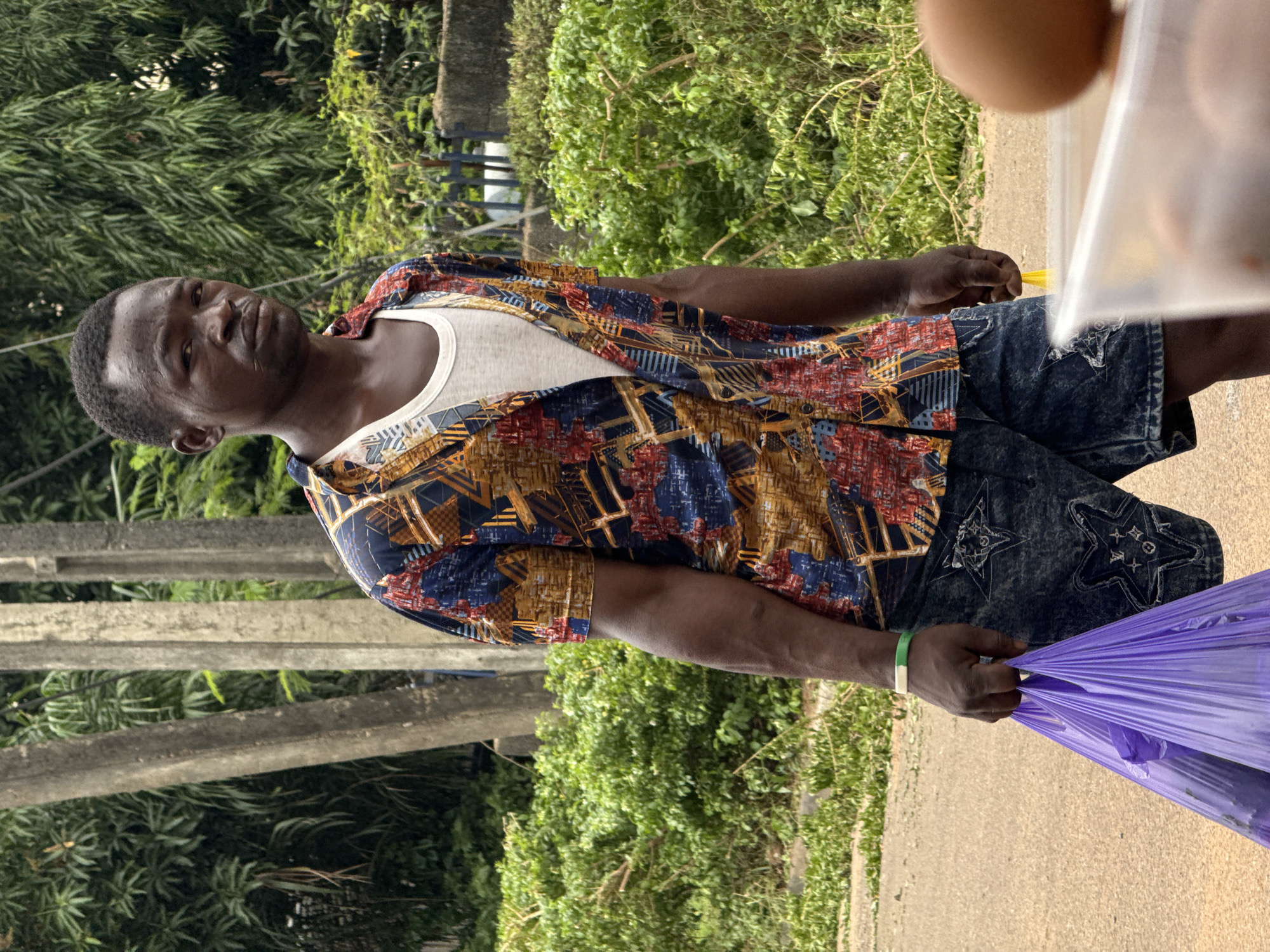 An outdoor, eye-level, medium shot captures a man in Abuja, Nigeria. He is standing on a dirt or gravel surface, holding a purple plastic bag in his left hand and a light brown object, possibly an egg, in his right hand, which is resting on a clear plastic container. The man wears a colorful, short-sleeved button-up shirt over a white tank top, and denim shorts with star-shaped patterns. A green and white bracelet is visible on his left wrist. The background features lush green foliage, trees, and what appear to be concrete structures. The lighting suggests it is daytime.