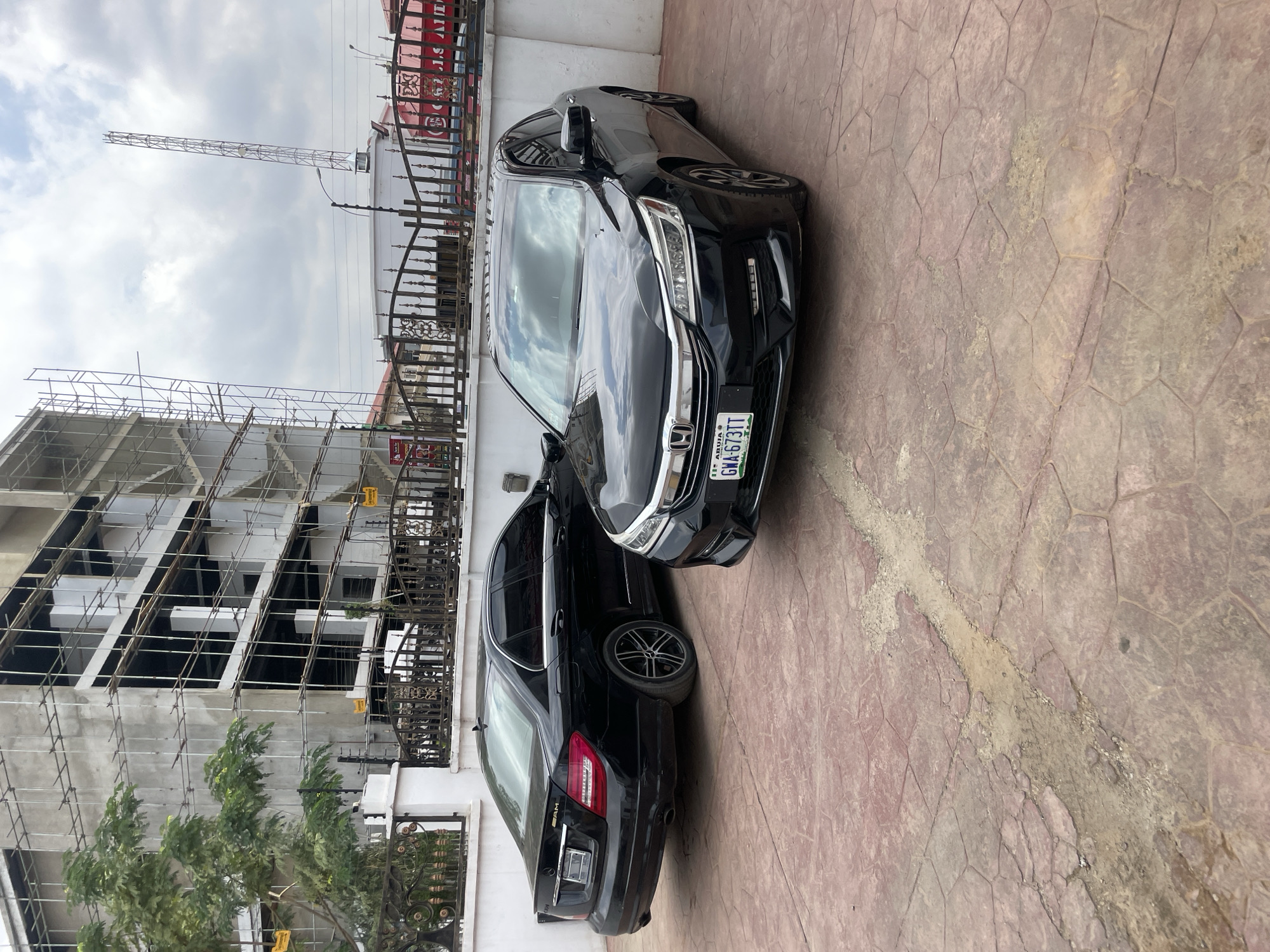 An outdoor, slightly rotated shot captures two black sedans parked side-by-side on a reddish-brown patterned pavement. The car in the foreground is a Honda Accord, recognizable by its grille and headlights, with a visible license plate. Behind it, a Mercedes-Benz C-Class (indicated by the "AMG" badge on the trunk) is partially visible.
In the background to the left, a multi-story building is under construction, enveloped in metal scaffolding. Further right, a decorative wrought-iron fence separates the parked cars from what appears to be another property or street. Beyond the fence, a tall, slender metal structure, possibly a telecommunications tower or billboard frame, rises against an overcast sky. Some green trees are visible near the construction site.
The ground features a distinct hexagonal or irregular tile pattern, showing some wear and lighter patches, possibly dirt or sand. The sky is largely covered with clouds, indicating daytime.
Visible text includes the license plate on the Honda Accord, which reads "ABUJA" and "GWA-673TT". The "AMG" badging is discernible on the trunk of the Mercedes-Benz. The setting suggests an urban environment in Abuja, Nigeria. There are no people visible, and no specific activity is taking place beyond the cars being parked.
