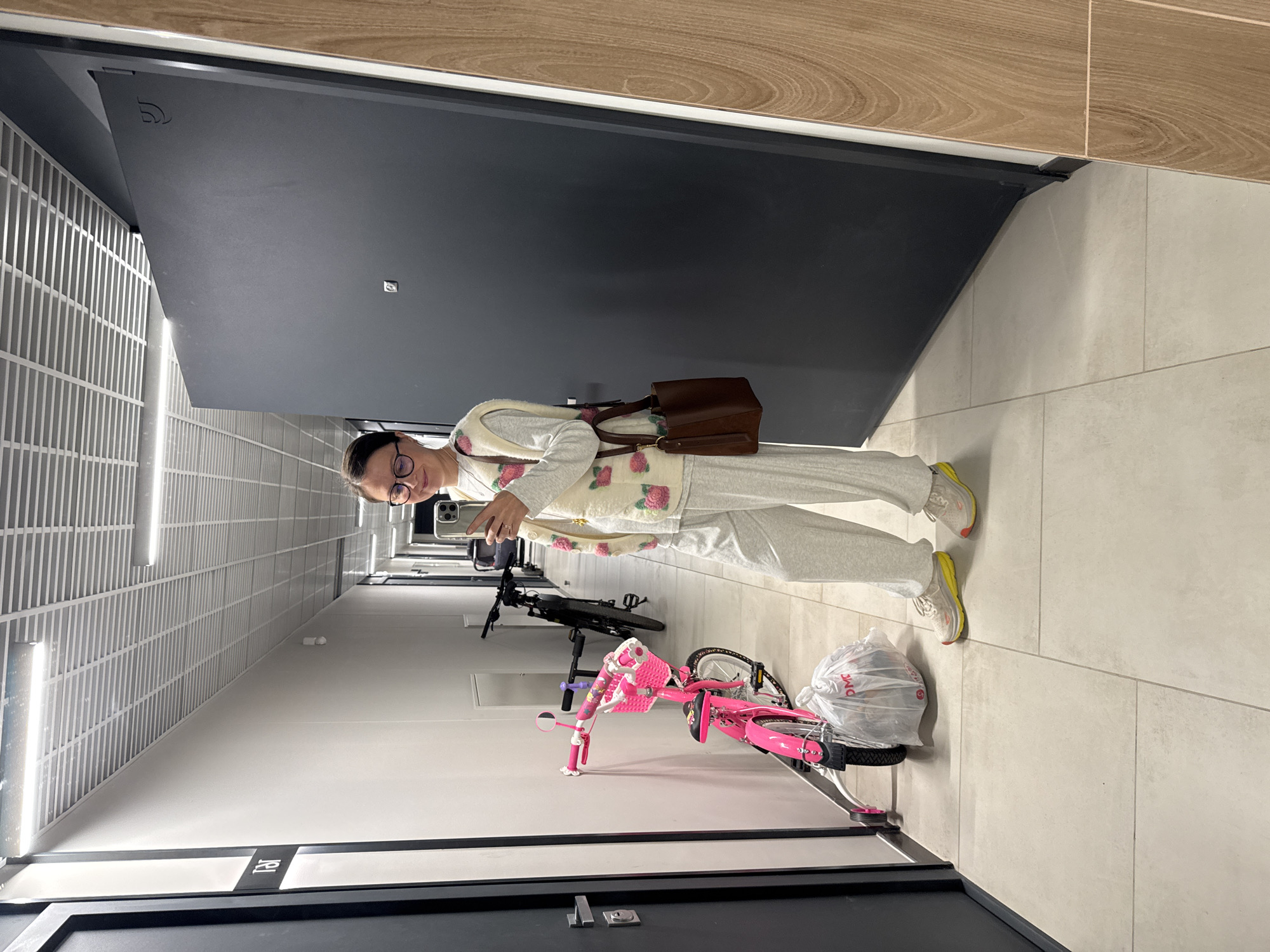 A woman takes a mirror selfie in a well-lit, modern indoor hallway in Izhevsk, Russia. She stands towards the center-right of the frame, holding a smartphone with her right hand, capturing her reflection. She is dressed in a casual outfit consisting of light grey sweatpants, a long-sleeved top, and a cream-colored sleeveless vest with pink rose embroidery. She wears eyeglasses and athletic shoes with yellow accents, and carries a brown leather shoulder bag.
The hallway features a light-tiled floor and walls, with dark, flat panel doors visible along both sides. One dark door behind the woman displays a stylized symbol resembling an intercom or doorbell, along with a small square fixture. Further down the corridor, a dark vertical strip on a wall or door is marked with the number "19". The ceiling is a gridded panel with bright, linear light fixtures.
Two bicycles are parked along the wall to the woman's left. The one in the foreground is a bright pink child's bicycle, complete with a front basket and a small handlebar mirror. A clear plastic bag, bearing the partial text "ДМС", rests on its rear wheel. Behind it, a black adult bicycle is also visible. The setting appears to be a clean, contemporary residential or commercial corridor.