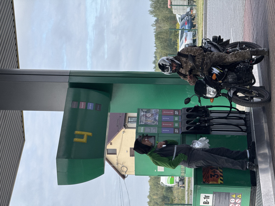 Two men are present at a green and grey fuel pump island located at a gas station in Starovoitove, Ukraine, on an overcast day.
To the left, a man stands beside the pump unit. He wears a dark grey hooded vest over a green long-sleeved shirt with a reflective stripe, and dark trousers. He holds a small, light object near his face and has a white plastic bag containing gloves attached to his left hip.
To the right, a second man is seated astride a black motorcycle. He is dressed in camouflage attire, including a jacket and trousers, and wears a white and black full-face helmet with a dark visor. He holds and is looking down at a smartphone with both hands. The black motorcycle has a front headlight, handlebars, and a visible exhaust pipe. A dark backpack is worn by the rider.
The fuel pump unit is predominantly green with a prominent yellow number "4" on its upper display. It offers multiple nozzles for "ДП" (Diesel) and "95" octane gasoline, labeled with "Mustang 95," "AVI Cappe 5," and "ДП Prime" on small screens. A poster with Ukrainian text is affixed to the pump. The base of the pump shows "B-1r" and hazard symbols.
In the background, behind the pump, a light-colored, possibly two-story, residential house with a dark brown roof and a security camera is visible. A line of green trees forms the horizon. To the far left, a portion of a white and green tanker truck is visible behind a fence. To the far right, various parked cars and a larger vehicle are discernible in a lot. The sky is cloudy.