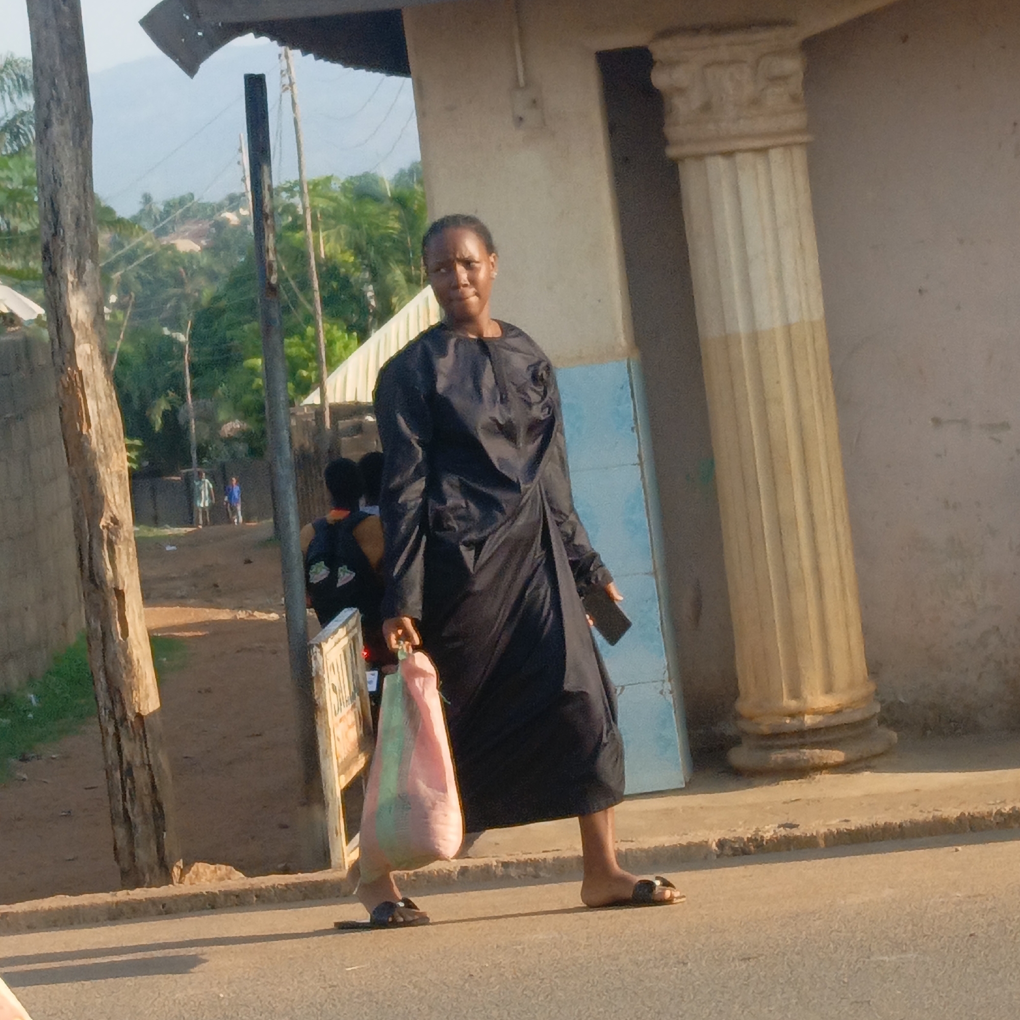 A medium shot captures a woman in Akwanga, Nigeria, standing on a paved road. She wears a long-sleeved black dress and sandals, carrying a full pink and green plastic shopping bag in her left hand and a cell phone in her right. Behind her, two people on a motor bike, partially obscured by the woman and what appears to be a "SALE" sign, are traveling away from the camera. In the distance, two figures are visible on a dirt road. The background features a building with a painted column and a blue-painted section. A utility pole is near the road and green vegetation and a mountain are visible in the distance.