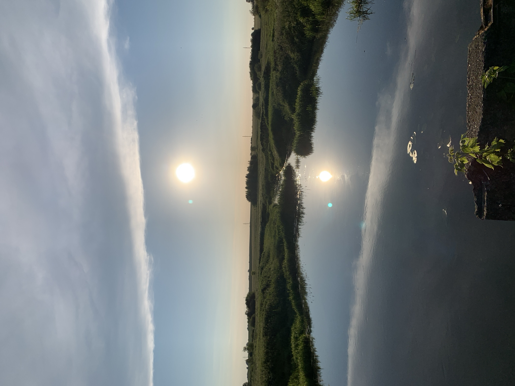 The image depicts a serene natural landscape at dusk, likely within the Блеўчыцкі сельскі Савет region of Belarus. A calm body of water occupies the foreground, reflecting the bright, low-hanging sun and the cloudy sky above. The sun appears as a luminous orb, slightly to the left of center in the sky, with its reflection visible on the water's surface. The water is bordered by lush green vegetation, including tall grasses and bushes, which extend into the middle ground of the image. In the distance, a flat, open field is visible, punctuated by a line of trees and faint silhouettes of utility poles, suggesting a rural or semi-rural setting. The overall atmosphere is peaceful and tranquil, characteristic of a summer evening in the Belarusian countryside. There are no people or animals visible in the scene, and no discernable actions or interactions are taking place. The focus is purely on the natural environment and the play of light on water and land.
