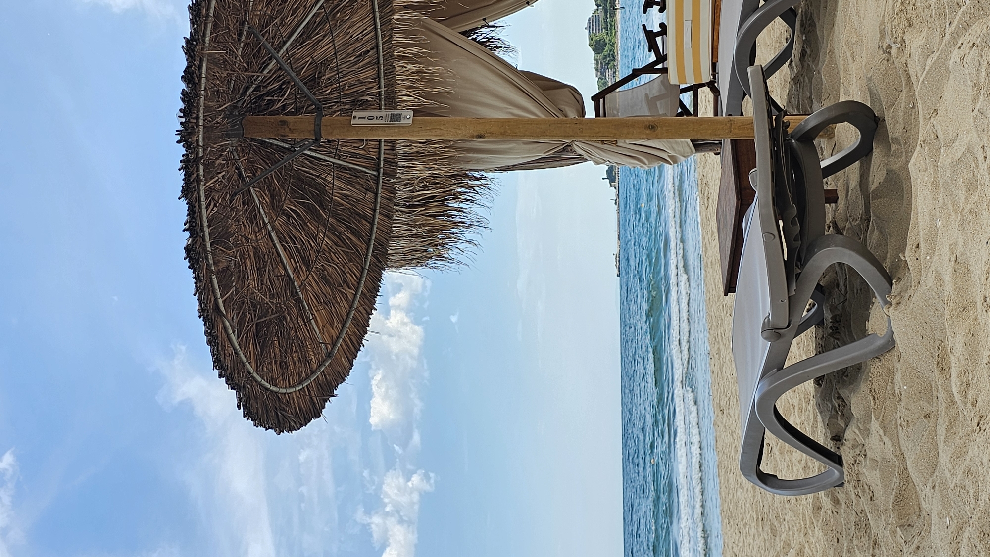 The image shows a beach scene in Varna, Bulgaria, oriented vertically. In the foreground, a large thatched umbrella casts a shadow over the sandy beach. Beside the umbrella, several grey plastic lounge chairs are arranged on the sand. To the left of the chairs, a wooden table is visible. The sea stretches out in the background, with gentle waves breaking on the shore. The sky is a clear blue with a few scattered white clouds. The overall atmosphere is peaceful and relaxing, suggesting a sunny day at the beach.
On the wooden pole of the thatched umbrella, a small silver tag with black text is visible. The text appears to read "105". The scene is likely set during daytime, given the bright sunlight and blue sky. The presence of lounge chairs and umbrellas indicates a tourist or recreational area. The calm sea and clear sky suggest pleasant weather conditions.
