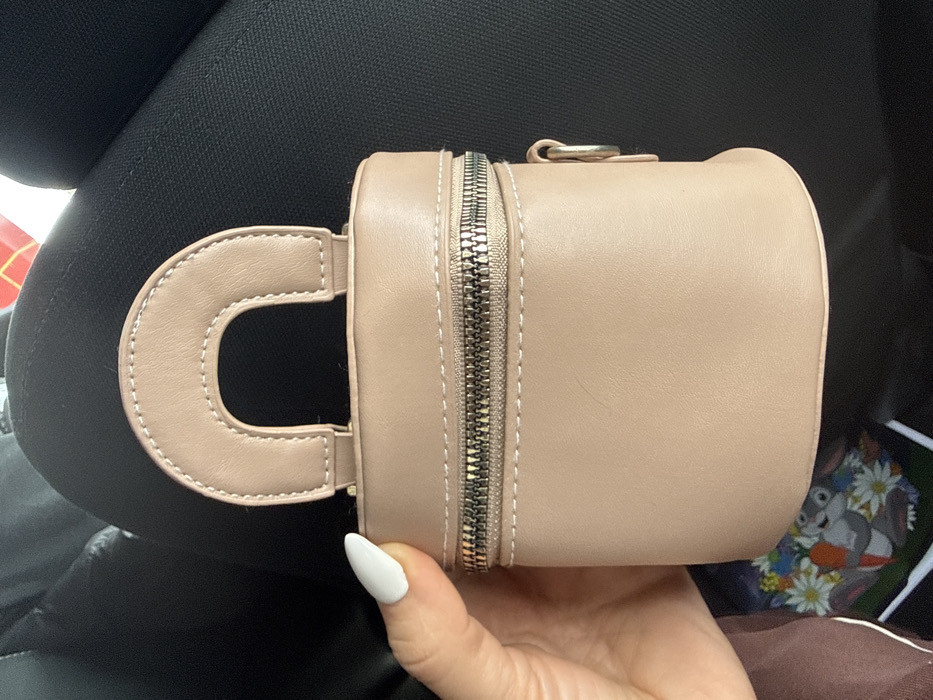 The scene depicts a light beige cylindrical handbag with a semi-circular top handle and a metallic zipper, held by a person's hand with white-painted fingernails. The handbag is positioned against a dark, textured background consistent with vehicle upholstery. Partially visible below the handbag is a colorful, blurred item featuring cartoon characters, including what resembles Bugs Bunny amidst carrots and daisies. A red and white object is subtly visible in the upper right background. The event is located in the city of Apnagyugh, Armenia.