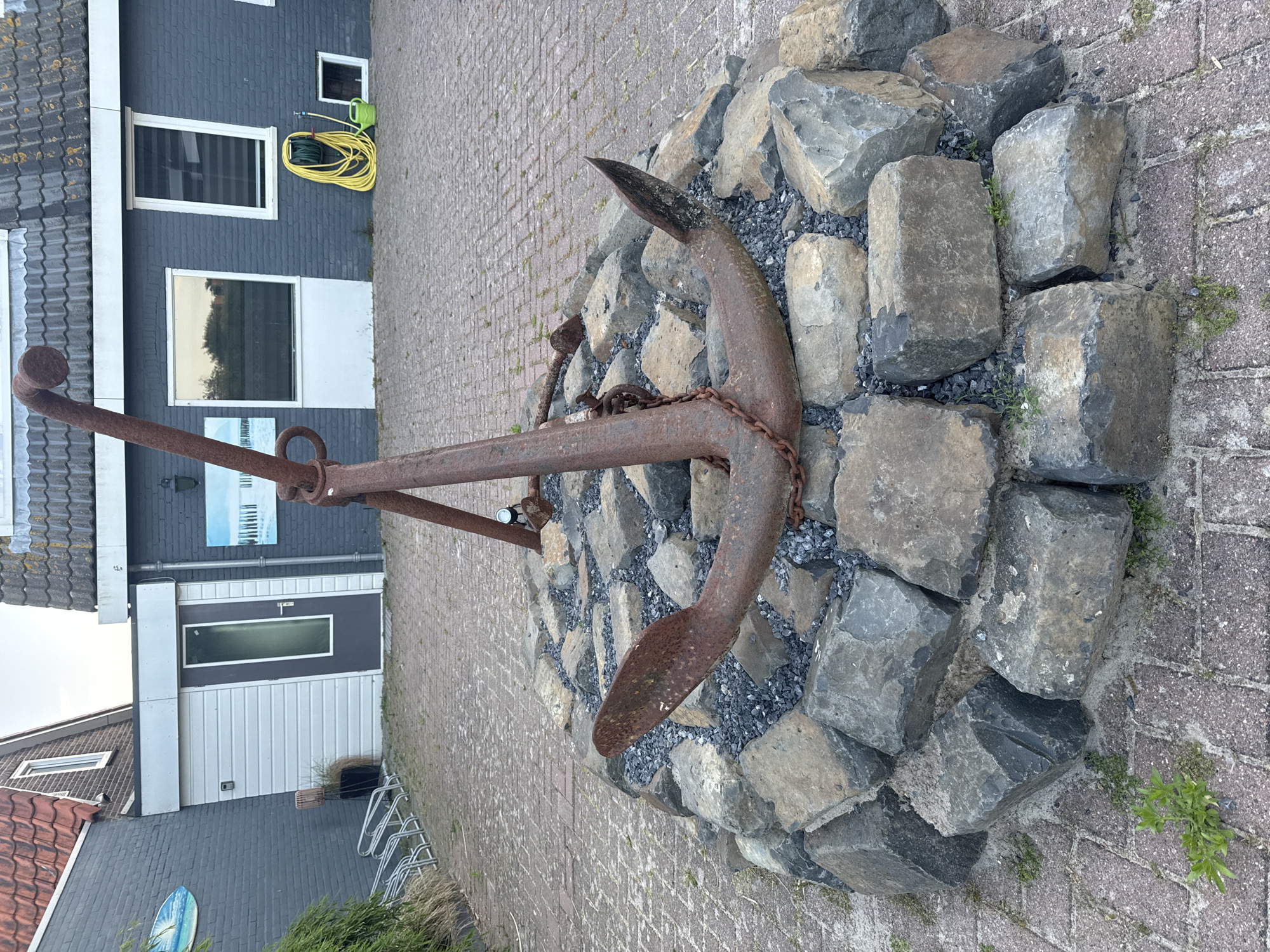 The image depicts an anchor monument in Petten, Netherlands. The rusty, weathered anchor is the central object, resting atop a circular base of large, roughly hewn gray stones. Small, dark gravel fills the spaces between the stones. The monument is positioned on a paved surface composed of interlocking bricks, with sparse vegetation growing along the edges.
In the background, a building with a dark gray facade and white trim is visible. It has two prominent windows and a door on the ground level. A coiled yellow garden hose hangs near one of the windows. The building is located on the edge of a street. A bike rack sits to the right of the door. A glimpse of additional buildings and structures can be seen in the lower part of the image, suggesting a typical Dutch village setting.