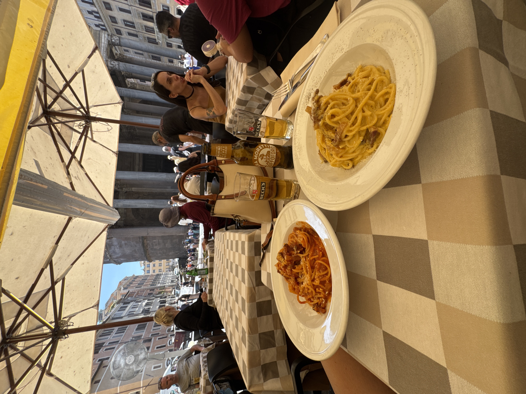 The image shows an outdoor dining scene. A person, presumably the photographer, holds a plate of spaghetti with red sauce at the bottom of the image, while another plate of spaghetti carbonara sits near the top. Two glasses of beer sit between the plates, with a bottle of Birra Moretti and a bottle of beer with a mostly obscured label. The table is covered with a brown and tan checkered tablecloth. A yellow umbrella provides shade from the sun. The background includes a crowd of people, buildings, and what appears to be the Pantheon in Rome, Italy. Several people are seated at the table. One woman in a black dress is reaching for a breadstick.