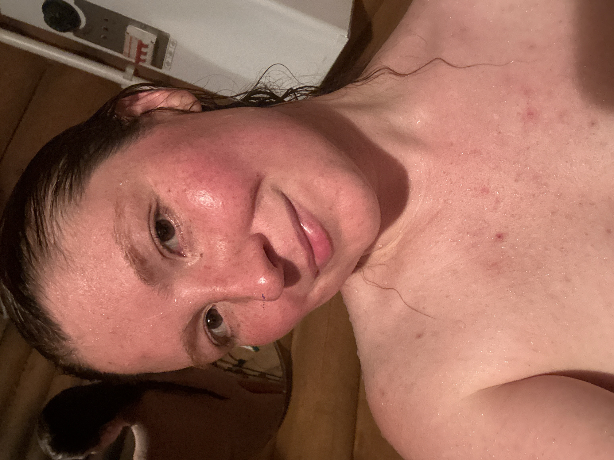 A close-up, slightly angled, portrait-style shot of a person with fair skin and wet hair. The person has visible pores, some redness, and a few blemishes on their face and chest. The person is looking at the camera with a slight smile. Water droplets are visible on their skin and hair, suggesting they may have just come out of a shower or bath. The background includes a beige or brown textured surface, which may be part of a bathtub or shower. There are also some indistinct objects and a white surface in the upper left corner. There's no evidence this image was taken in Nizhny Novgorod, Russia.