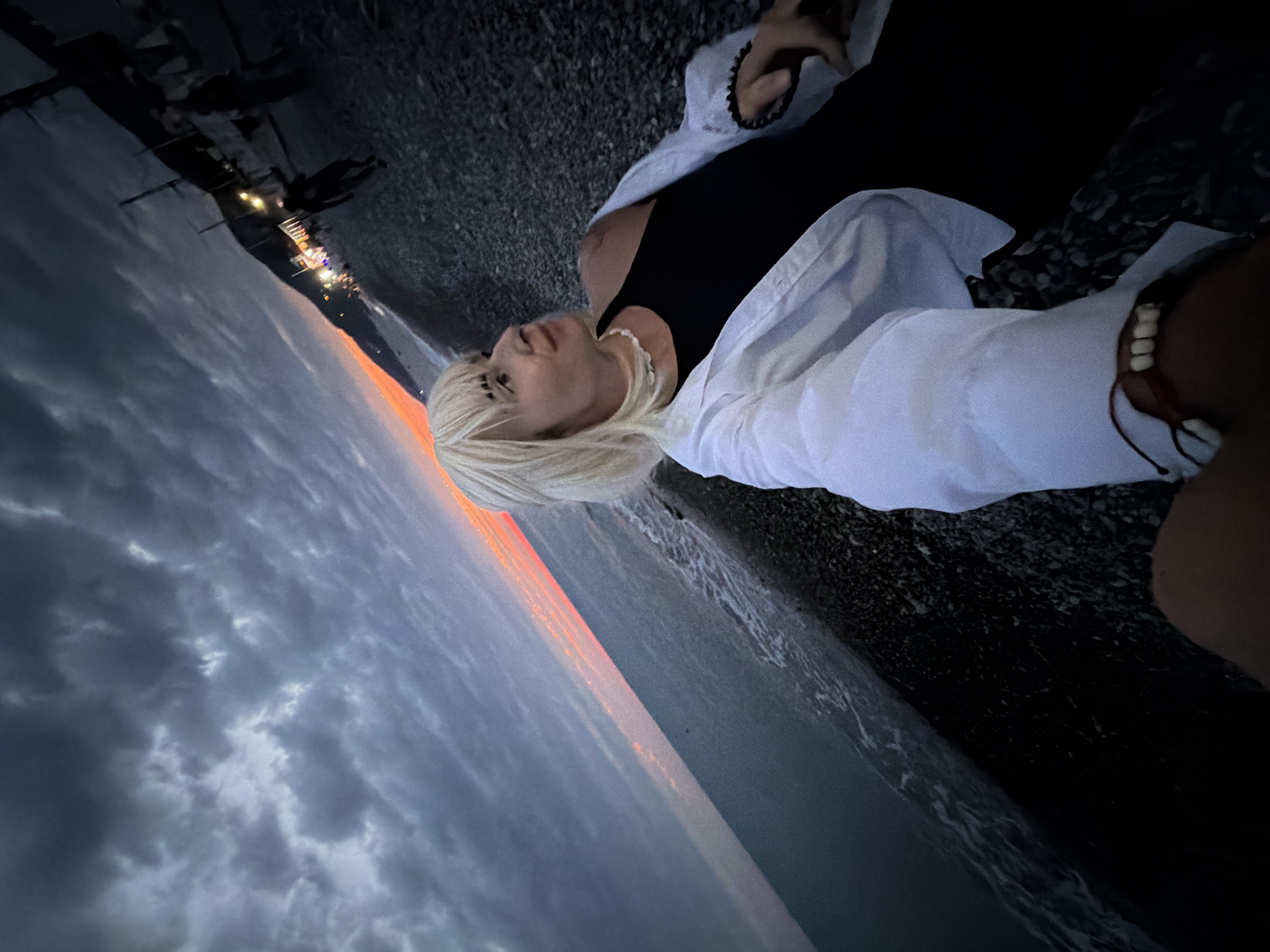 A person with blonde hair and dark makeup on their eyebrows is on a pebble beach at sunset, near the city of Sochi, Russia. The sky is dark with dramatic clouds, and a sliver of orange and pink sunset is visible on the horizon where the sky meets the sea. The person is wearing a white open shirt over a black top and a white beaded necklace. They are taking a selfie with their right hand, which is out of frame, and looking to the right, slightly away from the camera. The beach has dark pebbles, and the sea is calm with small waves. In the background, along the coastline, there are lights from buildings and possibly a promenade, and a few other silhouetted figures can be seen in the distance. The overall impression is of a serene, yet atmospheric evening scene.