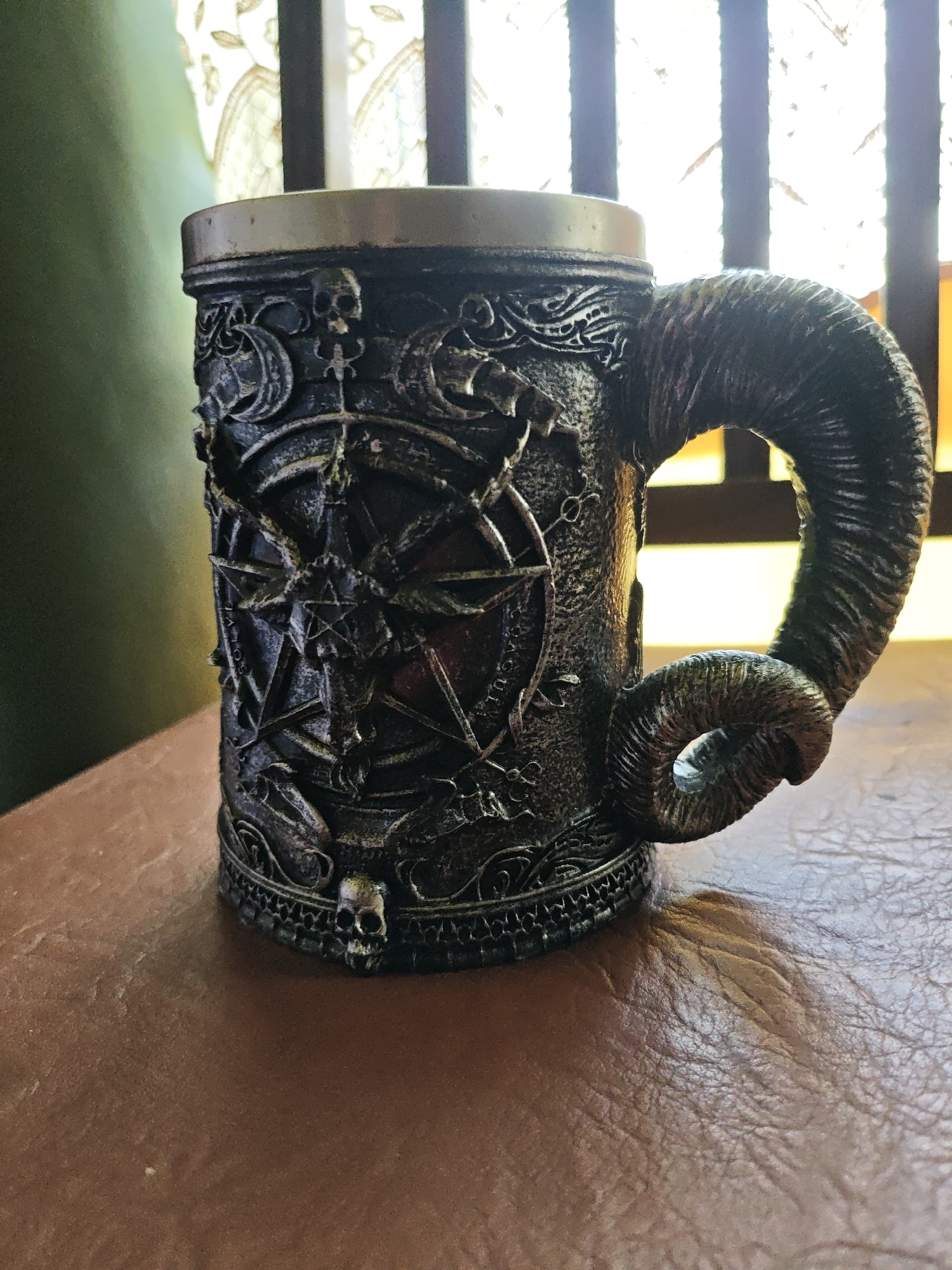 A close-up shot presents a gothic-themed mug. The mug is predominantly dark gray with ornate detailing, including skulls and a prominent pentagram. Its handle is shaped like a ram's horn. The mug sits on a textured brown surface. In the background, partially obscured, is a window with visible bars. There is no visible information to determine the city, or country it could be from.