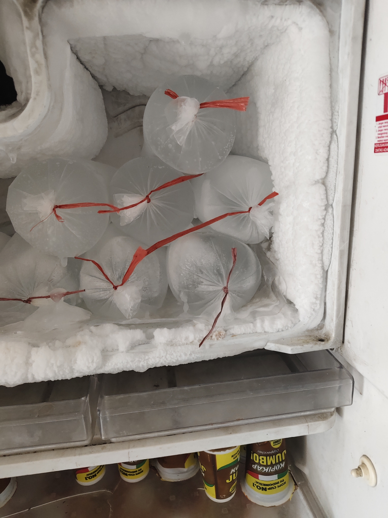 The image depicts the interior of a freezer, likely in a residential kitchen in Bangsalsari, Indonesia. The freezer is heavily frosted, with ice buildup visible on the walls and ceiling. Several clear plastic bags, each containing a frozen substance and tied with red string, are stacked inside. Below the bags, two clear plastic drawers are visible. Further down, on a shelf, are several cups of "Kopikap Jumbo" instant coffee, some of which appear to be partially used. The overall impression is one of a functional but somewhat cluttered freezer, typical of a household in an Indonesian city.