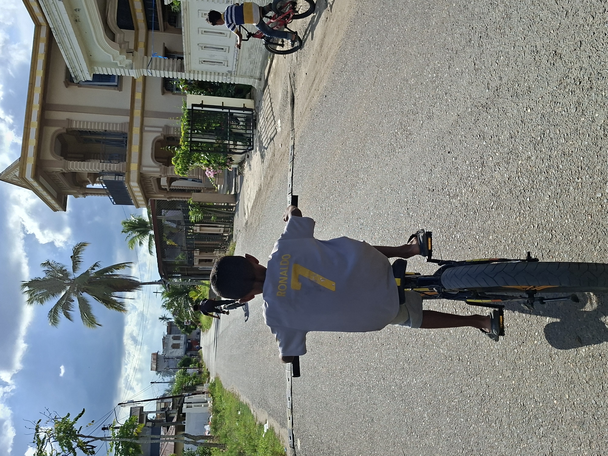 A sunny day in Lhokseumawe, Indonesia. A boy wearing a light-colored t-shirt with the number "7" and the name "RONALDO" on the back rides a bicycle down a paved road. He wears sandals. Another boy on a red bicycle is further ahead. Buildings, foliage, and a palm tree line the roadside. The sky is blue with scattered clouds.