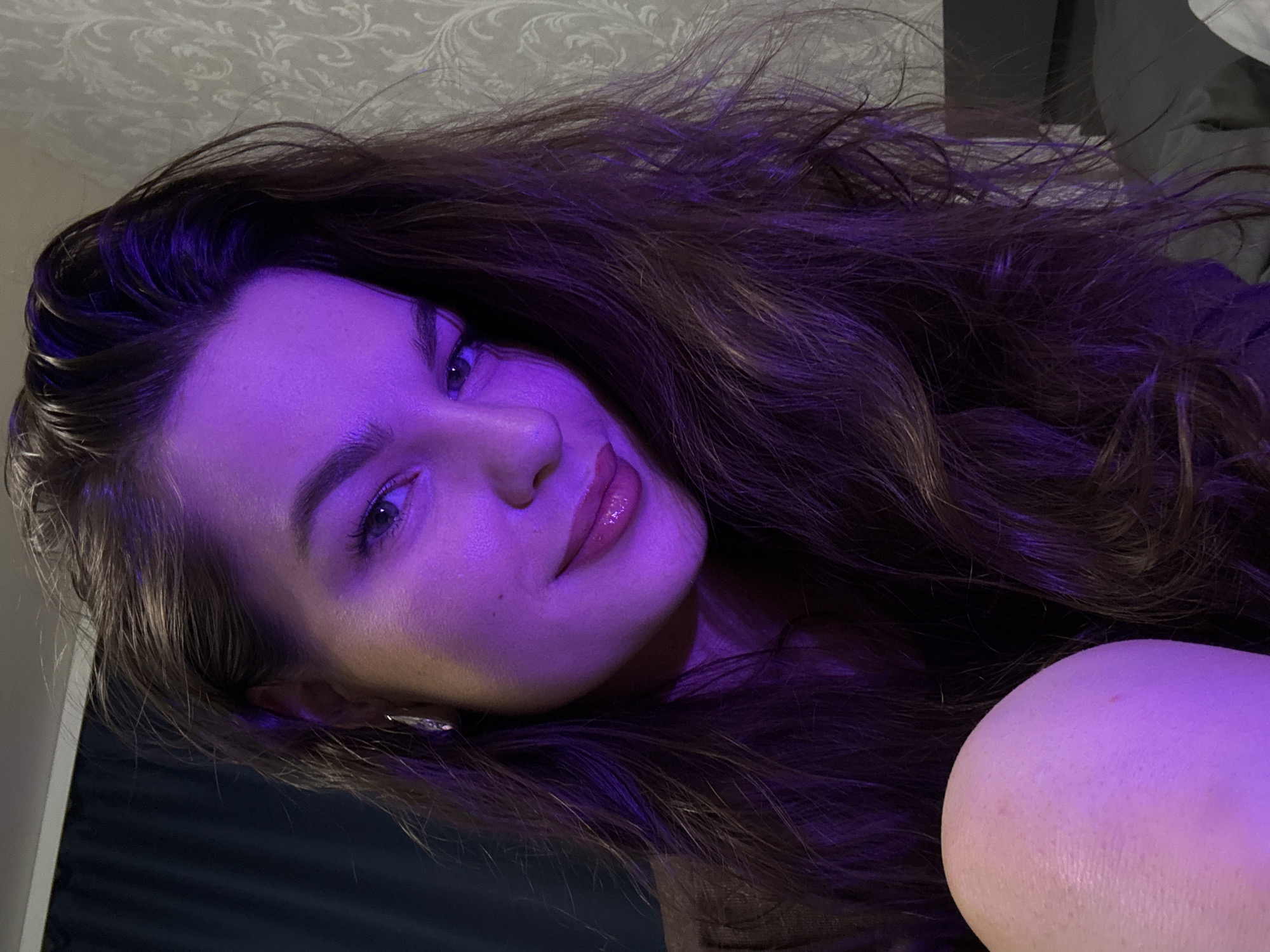 A woman is seen in a close-up, selfie-style shot, illuminated by purple lighting. She has long, dark, wavy hair that falls around her face and shoulders. Her left ear is adorned with a silver earring. She is wearing makeup, including lipstick that appears glossy. Her skin is smooth and fair, with a slight texture visible in some areas. The background is dimly lit and consists of a textured wallpaper on the right and a dark curtain or fabric on the left. A hint of her shoulder or arm is visible in the lower left corner of the frame, appearing smooth and illuminated by the same purple light. The overall environment suggests an indoor setting, possibly a bedroom or living space. The image does not contain any elements that would allow for a specific location within Chaikovsky, Russia.