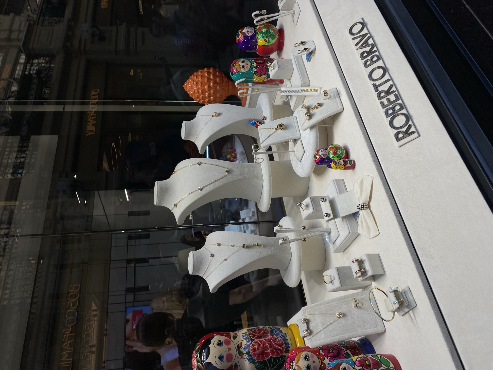 The image shows a jewelry display in a storefront window, likely in Moscow, Russia. The name "ROBERTO BRAVO" is visible on the display. The jewelry is presented on white stands and includes necklaces, earrings, and bracelets. Interspersed with the jewelry are decorative Russian nesting dolls (Matryoshka dolls) and an orange decorative object. The storefront window reflects the surrounding city buildings and other signs, possibly reading "ROCCO". People are visible as reflections in the glass.