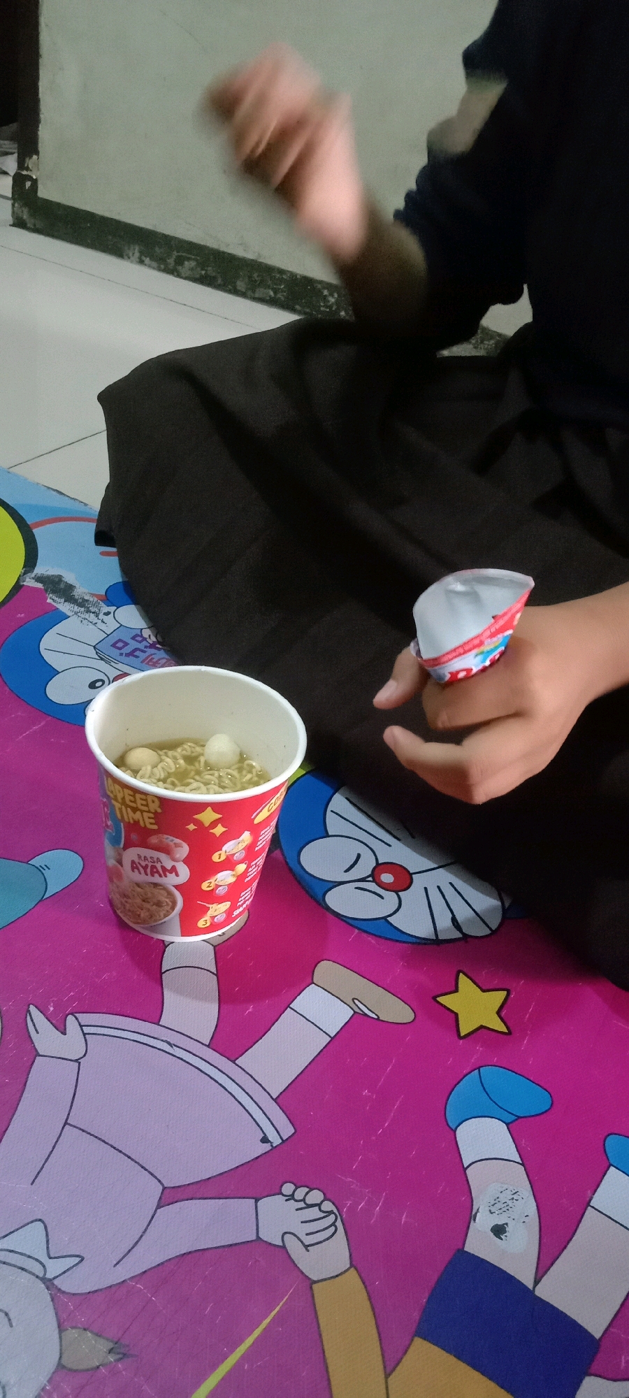 The image shows a close-up of a person's hands and a cup of instant noodles. The person is wearing a dark long-sleeved shirt and possibly a dark skirt or pants. One hand is blurred, indicating movement, and appears to be reaching towards the noodles. The other hand is holding a colorful, crushed cup, possibly a lid or an accessory from a food item, and is positioned near the cup of noodles. The cup of noodles is red and white with text and graphics, including the words "RASA AYAM" (chicken flavor). The noodles appear to be soup-based with some meatballs or dumplings. The setting is indoors, with a patterned mat featuring cartoon characters, including Doraemon, and other figures. The mat is on a tiled floor. The wall in the background is light-colored and appears somewhat worn. The scene suggests a casual, perhaps domestic, environment where someone is preparing to eat or has just finished eating instant noodles. The image does not contain any visual information that would allow for the identification of the city of Tasikmalaya, Indonesia, or any specific location within it.