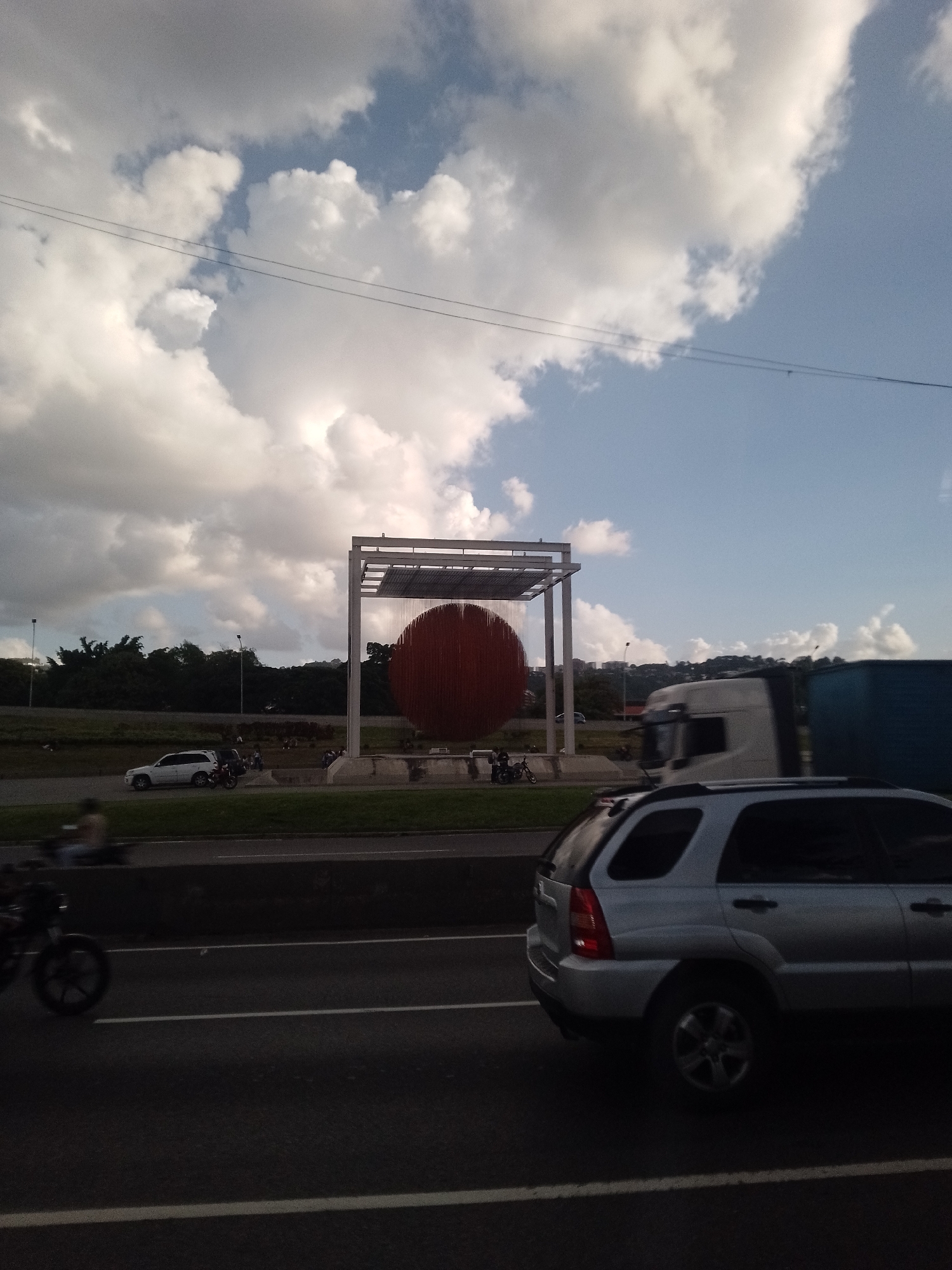 The image presents a dynamic scene on a multi-lane highway in Caracas, Venezuela, featuring the prominent "Esfera Caracas" sculpture by Jesús Rafael Soto.
In the mid-ground, the iconic kinetic sculpture, a large red spherical artwork composed of many individual elements, is housed within a minimalist white metallic rectangular frame. It stands on a raised platform, drawing the eye as a focal point amidst the surrounding traffic and greenery.
The foreground is dominated by vehicular movement. A silver SUV is partially visible on the right, while a blurred motorcycle and rider appear on the far left. On the highway lanes closer to the monument, several vehicles are present, including a white SUV and other motorcycles. A large white and blue semi-truck is in motion on the right side of the road, contributing to the sense of a busy thoroughfare.
The scene is set under a partly cloudy sky, with large, puffy white cumulus clouds dominating the upper portion of the frame, interspersed with patches of clear blue. Two thin power lines stretch horizontally across the sky. Lush green trees and foliage line the highway and form a dense natural backdrop, suggesting the tropical environment of Caracas.
The lighting indicates either late morning or late afternoon, characterized by soft, diffused daylight rather than harsh midday sun. The overall impression is of an active urban landscape, with the distinctive artwork serving as a significant landmark. No discernible text is visible in the image.