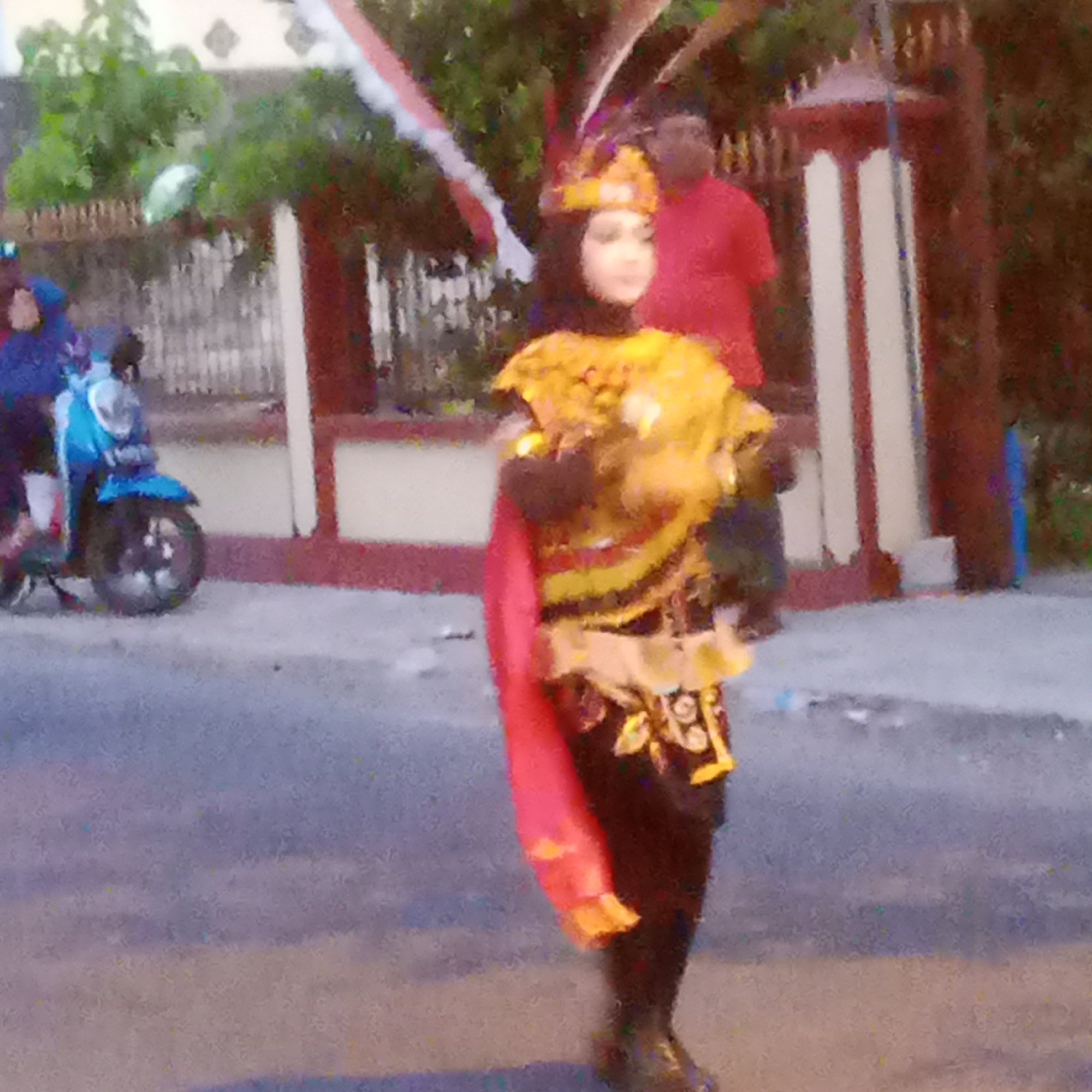 In the city of Semanding, Indonesia, a person in a colorful, traditional costume with a feathered headdress and gold accents is captured mid-stride on a paved street. The costume includes a yellow top with intricate patterns, a red sash draped over the left shoulder, and dark pants. The individual wears dark shoes. To the left of the person, a bright blue scooter is parked. Behind the scooter, other individuals are partially visible, dressed in casual clothing, suggesting a public gathering or event. In the background, a white fence with a red-brown railing runs along the street, with lush green trees and foliage behind it. A building with white columns and red trim is also present. The overall scene suggests a festive or cultural celebration taking place outdoors.